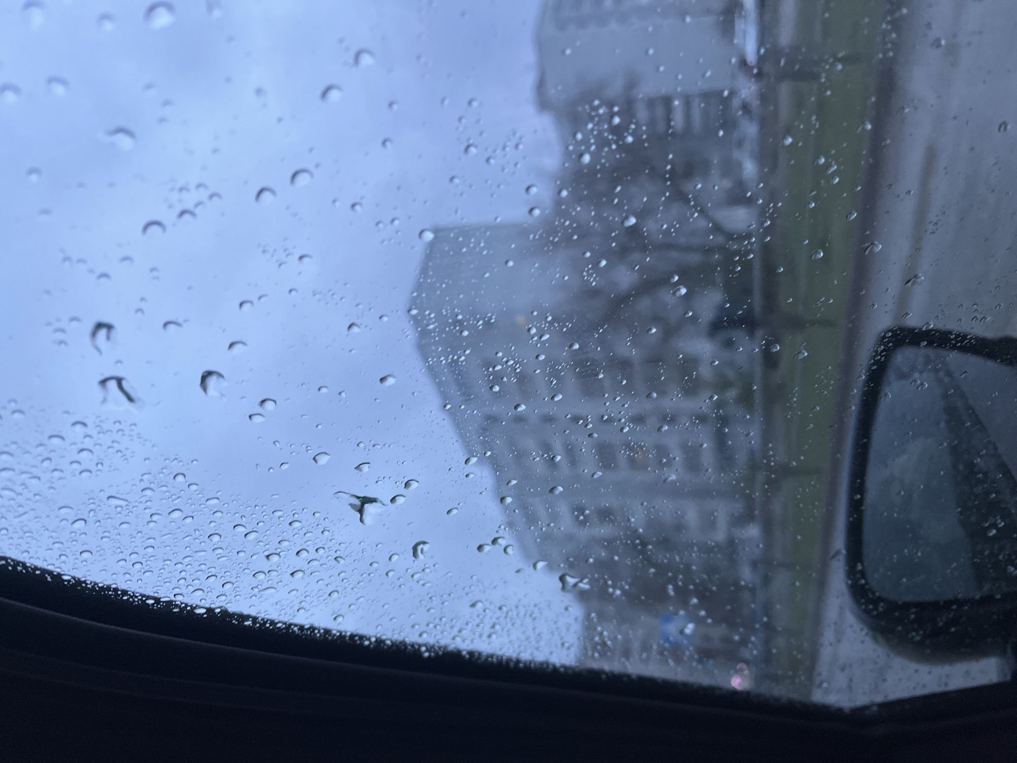 The image shows a rainy scene viewed through a vehicle's sunroof. Water droplets cover the glass. Outside, a blurred, tall building is visible, along with a car's side mirror on the right. The dark interior of the vehicle frames the bottom of the image. While the city is claimed to be Chaikovsky, Russia, it is difficult to confirm solely based on the provided information.