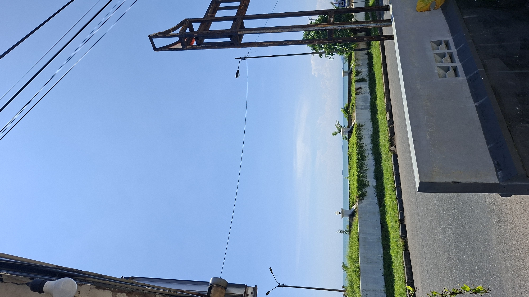 The image displays an outdoor scene under a clear blue sky, suggesting a sunny day. The perspective is from street level, looking upwards and outwards towards a body of water, likely the ocean, in the distance.

In the foreground, a concrete structure with square cutouts and a smooth, grey finish runs along the right side of the frame. Adjacent to this structure is an asphalt road with a dark curb. A strip of vibrant green grass borders the road and a concrete wall, separating it from a further section of manicured grass and foliage. Beyond this, the vast expanse of the ocean stretches to the horizon, meeting a pale sky with faint white clouds.

On the left side of the image, a series of black electrical wires are strung across the sky, originating from a dark fixture at the bottom left. A rusted metal frame, possibly part of a sign or construction, is visible in the upper left quadrant of the image. A street lamp with a single black bulb hangs from a wire extending from this frame.

The scene appears to be a public space, possibly a promenade or a street alongside a beach. The presence of the road, street lamp, and manicured green spaces suggests an urban or developed coastal area. There are no people visible in the image, and the overall impression is one of a quiet, possibly tranquil, environment. The lighting indicates it is likely daytime, with the sun casting shadows that are not prominently visible in this orientation. No text is discernible in the image.