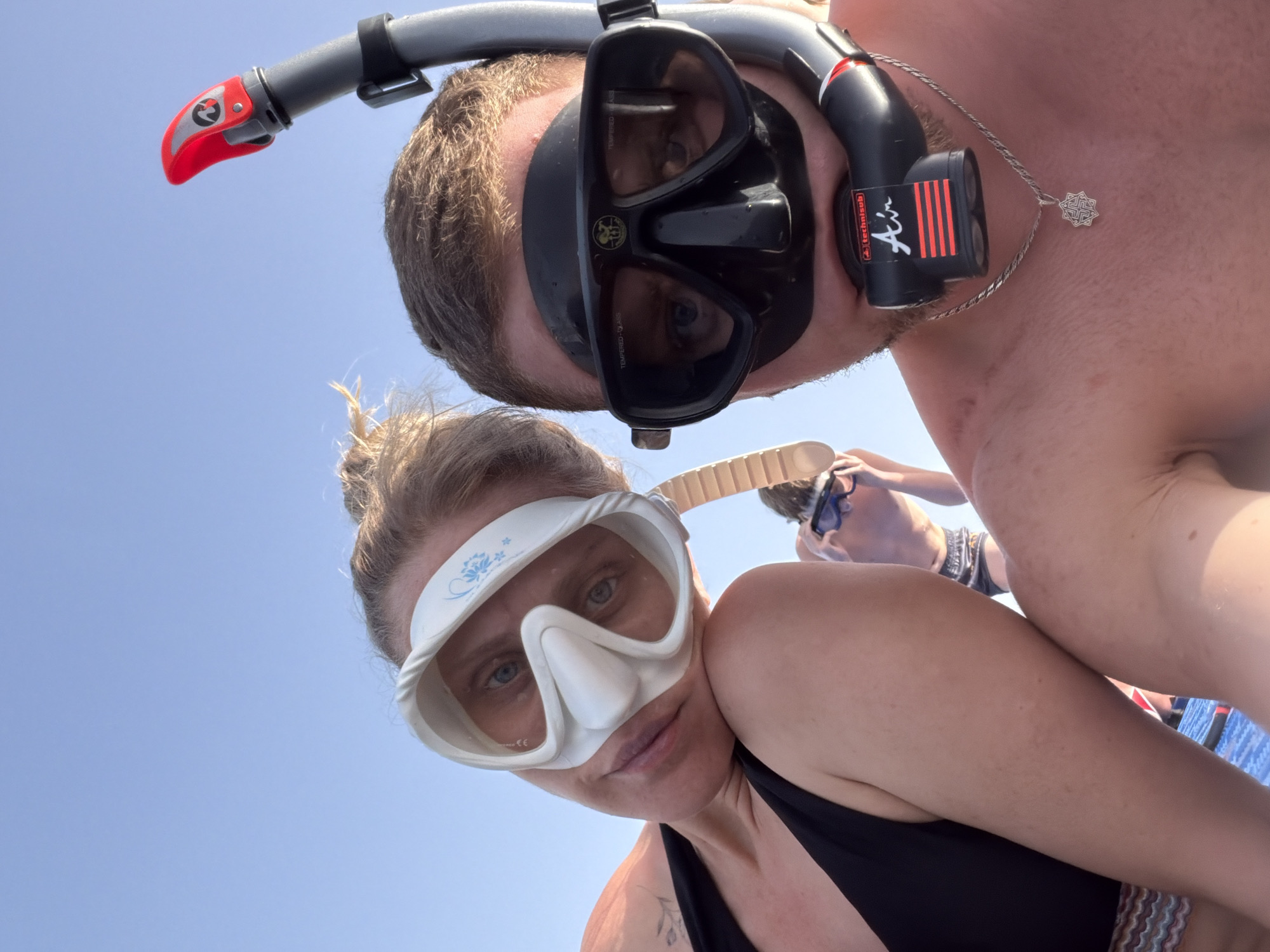 The image shows two people wearing snorkeling gear, viewed from a low angle looking up at them against a bright, cloudless sky. The person in the foreground is wearing a white snorkel mask and a black swimsuit. The person in the background is wearing a black snorkel mask and snorkel. Another person is visible in the distance. The image location is likely near a body of water, possibly in Sharm El Sheikh, Egypt.