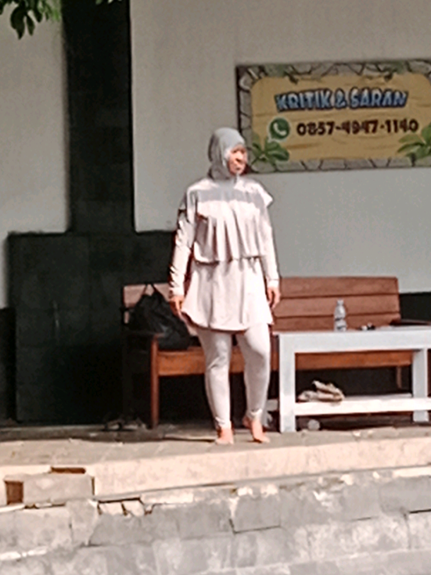 A woman wearing a grey hijab and a light pink tunic and pants stands on a concrete path. Behind her, a wooden bench is positioned in front of a wall. A white table with a wooden shelf underneath is to the right of the bench. A bottle of water and other small objects are on the table. A sign with the words "KRITIK & SARAN" and a phone number is mounted on the wall above the bench. The setting appears to be outdoors, possibly in a public space or a residential area in Jentekan, Indonesia, given the context of the sign which is typical for local businesses or community centers. The lighting suggests it is daytime.