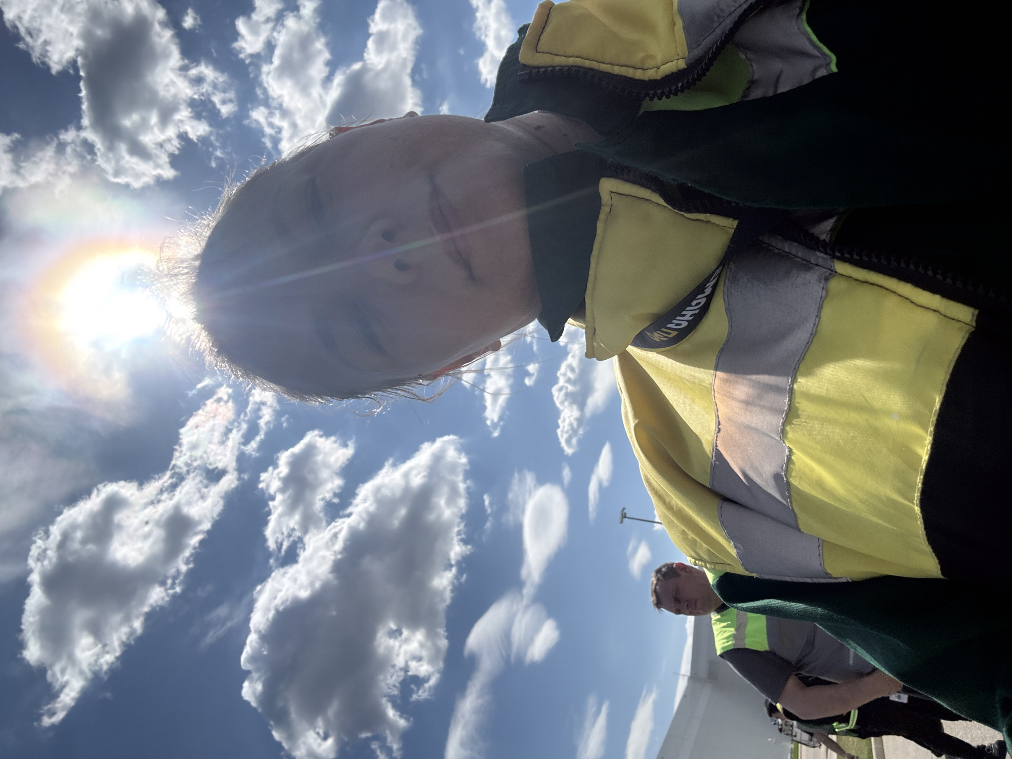 The image shows a selfie taken outdoors under a bright blue sky with puffy white clouds. The sun is directly behind the head of the person taking the selfie, causing a lens flare. The person taking the selfie is wearing a bright yellow and gray reflective vest over a black jacket. A portion of another person wearing a similar vest can be seen in the lower right corner of the image. The setting appears to be in a public space with light colored architecture in the background. The location cannot be determined with certainty. The city of Lupanovo, Russia is not shown in this image.