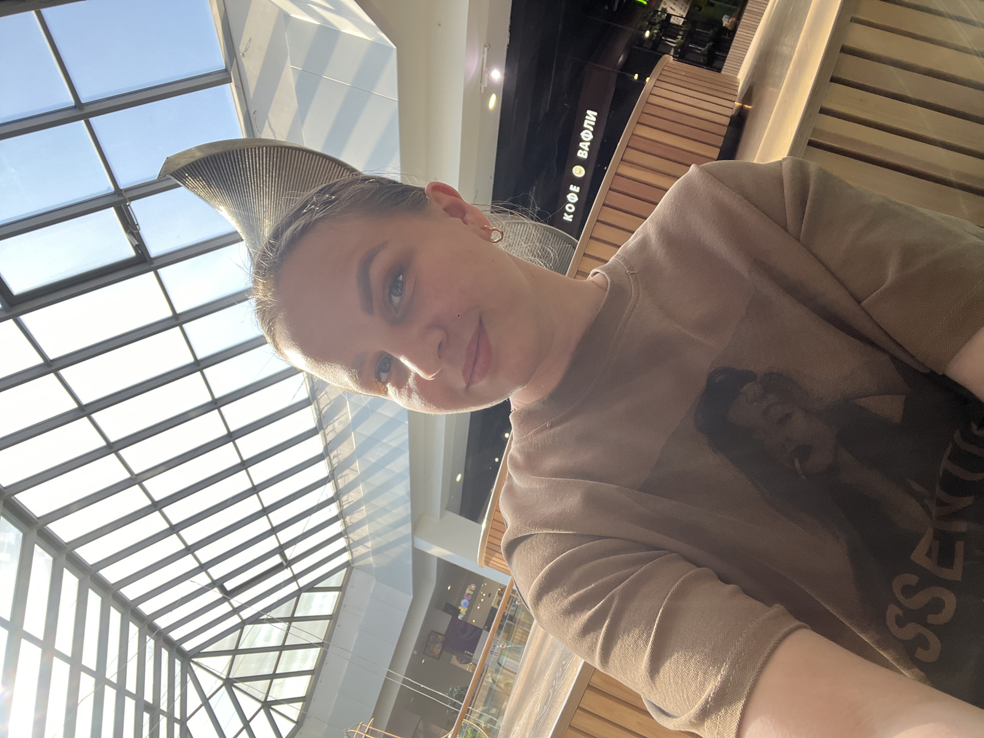 The image depicts a person seated indoors, likely within a shopping mall or similar public space. The person has fair skin and is wearing a brown t-shirt with a dark graphic. The environment includes a high, glass-paneled ceiling with metal supports, indicating a large indoor space. Wooden railings are visible, suggesting the person is on an elevated level. In the background, there is a sign in Russian "КОФЕ ВАФЛИ" ("Coffee Waffles"), indicating a cafe or food vendor. The city is identified as Izhevsk, Russia.