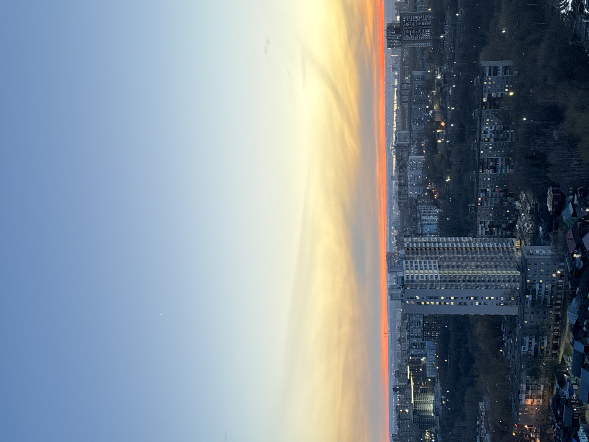 The image shows a sunset over Perm, Russia. The sky transitions from a light blue on the left to yellow and orange near the horizon. The city skyline is visible on the right, featuring numerous high-rise buildings, many with illuminated windows. There are dark forested areas interspersed among the buildings. The photograph is oriented vertically.