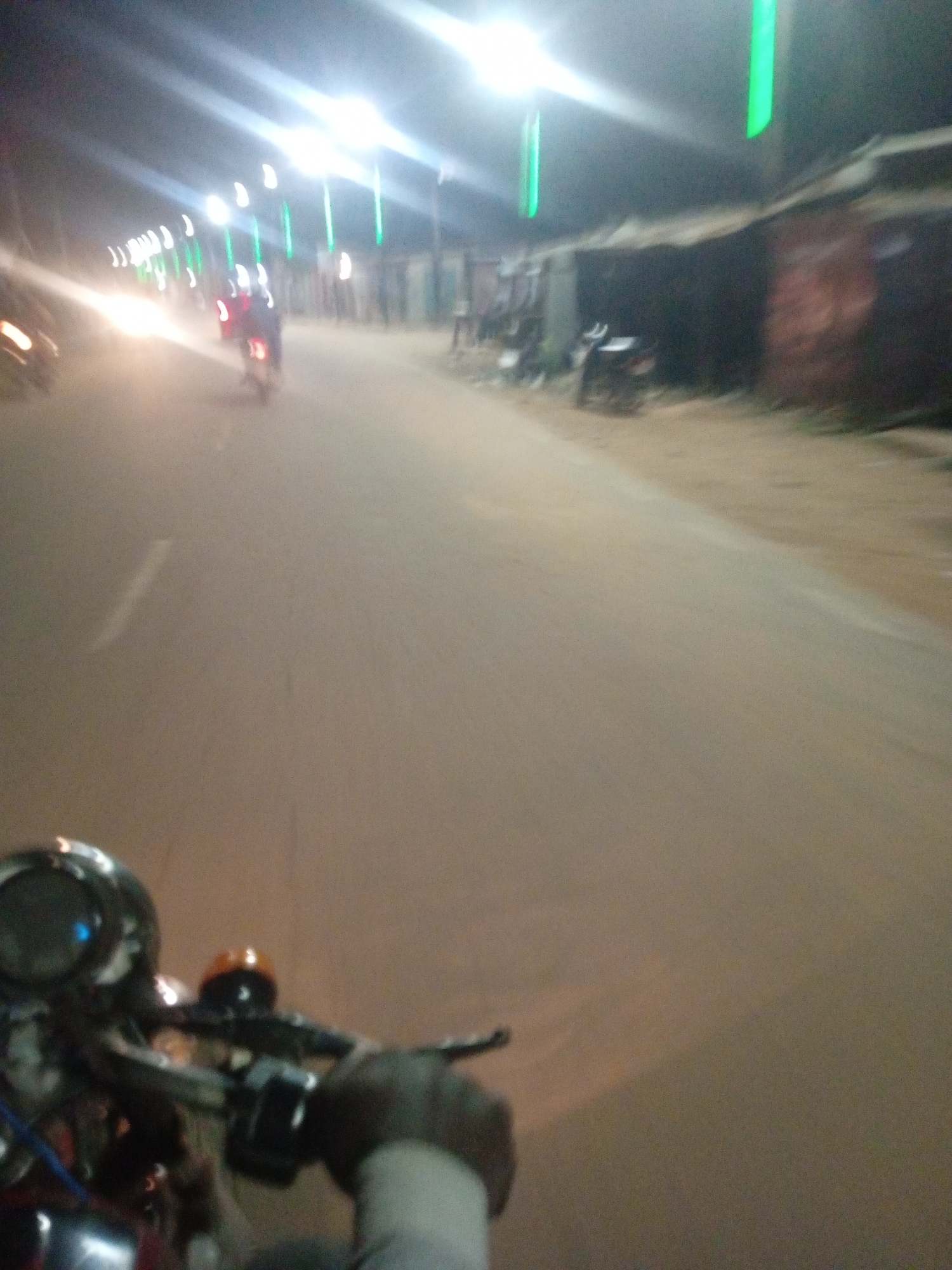 This image shows a nighttime scene on a street in Tundun Wada West, Nigeria. The perspective is from someone riding a motorcycle, with the handlebars and part of the dashboard visible in the foreground. The road appears to be paved with a sandy or dirt shoulder. Along the right side of the road, there are low-rise buildings with dark, possibly tin roofs. Several streetlights are illuminated, casting bright white light, and there are also some vertical green lights visible, possibly decorations or additional lighting.
In the distance, there are other vehicles, including at least one motorcycle with a rider. The lighting suggests it is evening or night, and there are no immediate cues about the weather other than the darkness. The overall impression is of a busy, illuminated street at night, perhaps during a local event or simply a regularly lit thoroughfare. The motion blur indicates movement, consistent with being on a moving motorcycle. No text is visible in the image. The scene is somewhat blurry due to the low light conditions and the movement.