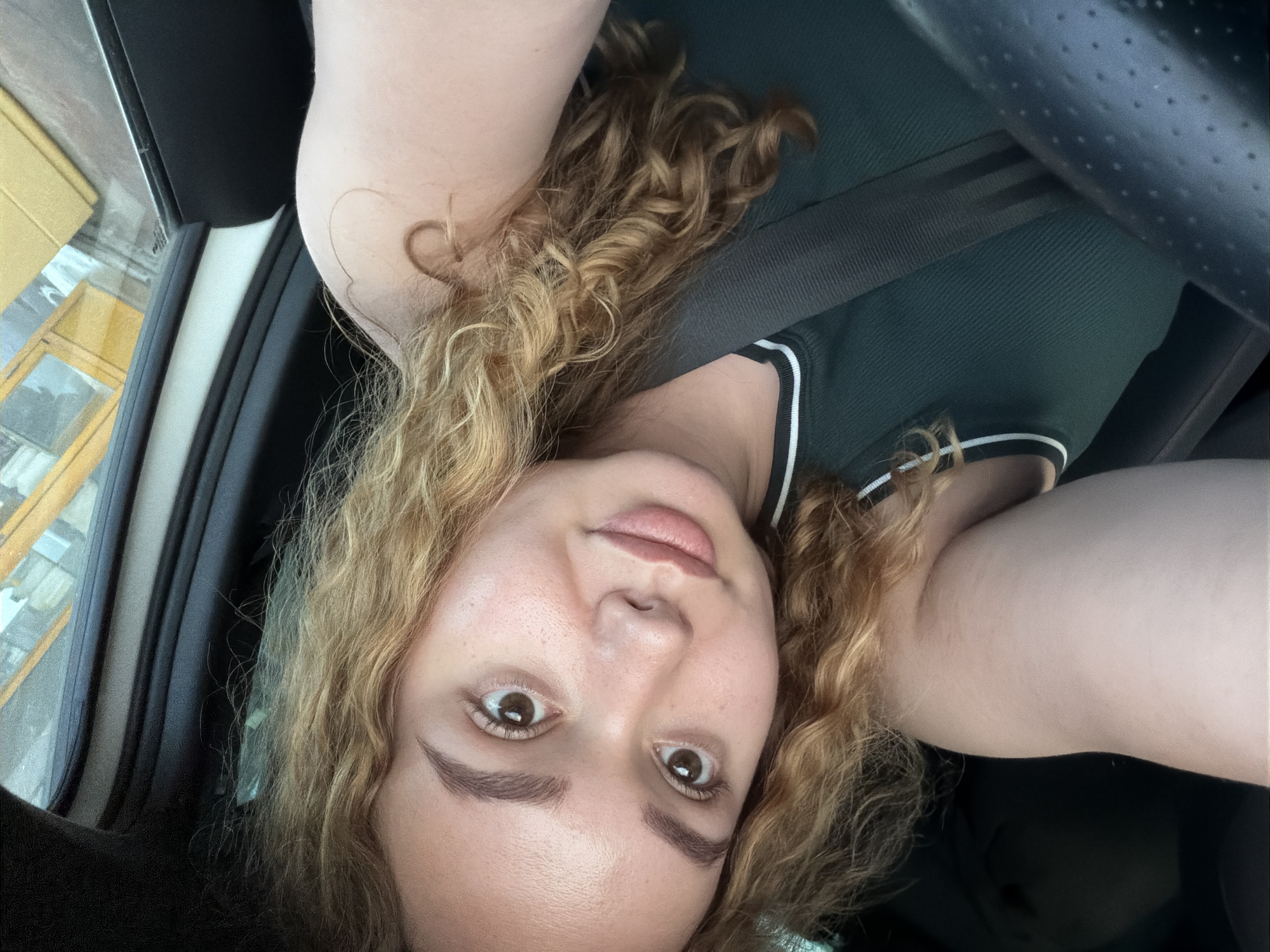 A close-up selfie, originally rotated 90 degrees clockwise, features a young woman inside a vehicle during the daytime. She has fair skin, striking brown eyes, full lips, and well-defined eyebrows. Her long, voluminous, curly hair, a blend of blonde and light brown, cascades around her face. A notable detail is a heart-shaped curl of hair near her left temple. She wears a dark green or teal sleeveless top with white stripes accentuating the trim.
The setting is clearly a car interior, evidenced by a black seatbelt diagonally crossing her chest. Parts of the vehicle's interior are visible, including a dark steering wheel on the right side and a perforated ceiling panel in the upper right corner. Through the window on the left, a building with yellow or tan siding and dark windows can be seen, suggesting an urban or suburban outdoor environment. The lighting implies it is daytime. The woman has a neutral expression, looking directly into the camera. There is no visible text.