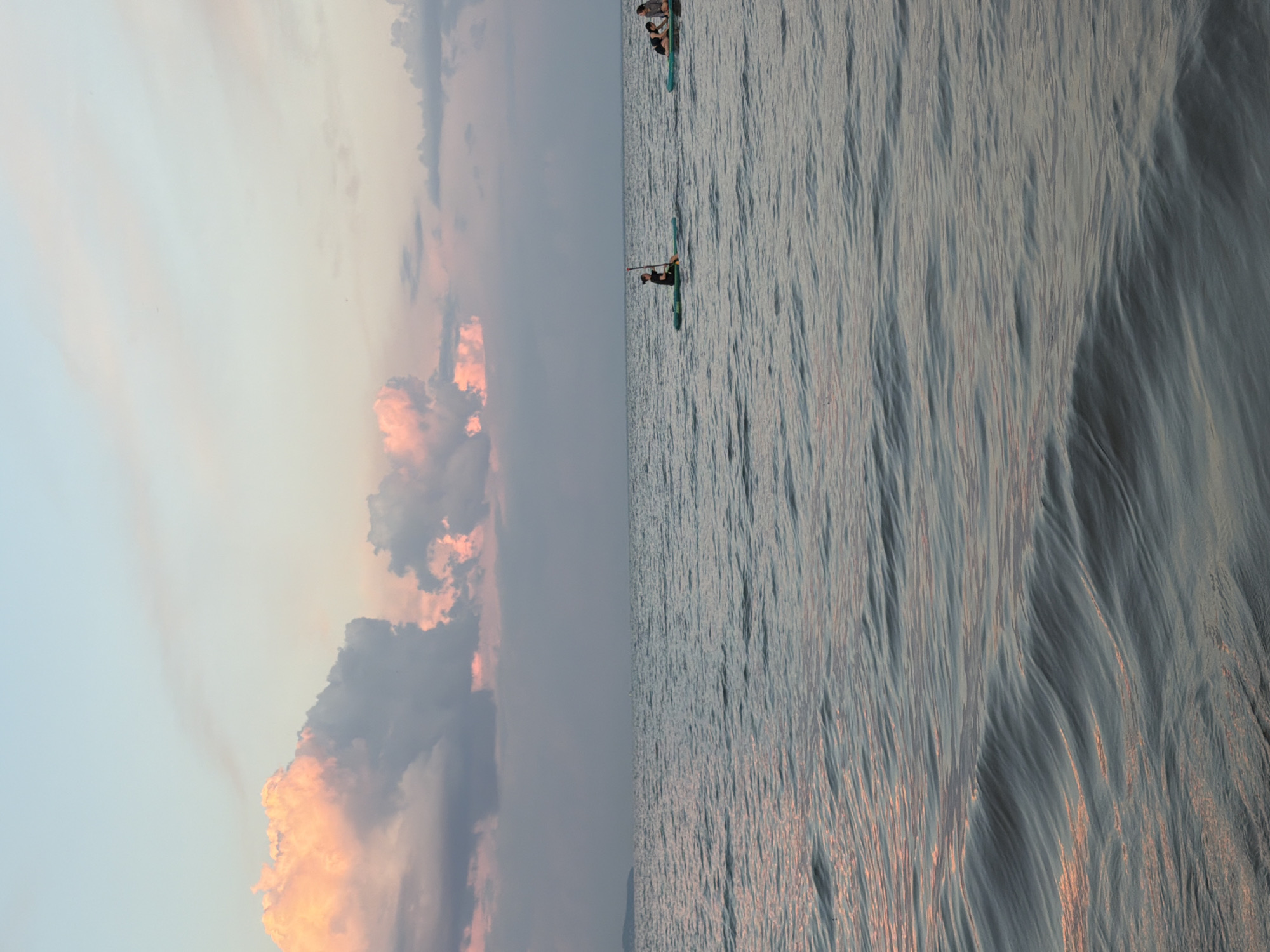 This is a vertical, eye-level medium shot of a serene seascape with a dramatic sunset sky. The image is split vertically, with the left side dominated by soft, pastel-colored clouds in shades of pink, orange, and blue, indicative of a sunset. The right side of the image features the calm, rippling surface of the water, reflecting the warm tones of the sky.
Several people are visible on paddleboards in the distance, engaging in a leisurely water activity. One person in the foreground is paddling a green paddleboard, while another group of two or three individuals is further back on a similar board. Their silhouettes are small against the vastness of the water and sky, suggesting a peaceful and tranquil atmosphere.
The time of day is clearly sunset, as indicated by the colorful sky and the soft lighting on the water. The weather appears calm, with gentle ripples on the water's surface and no visible harsh elements in the sky. The scene evokes a sense of peace and natural beauty. The location is Laishevo, Russia. There is no visible text in the image.