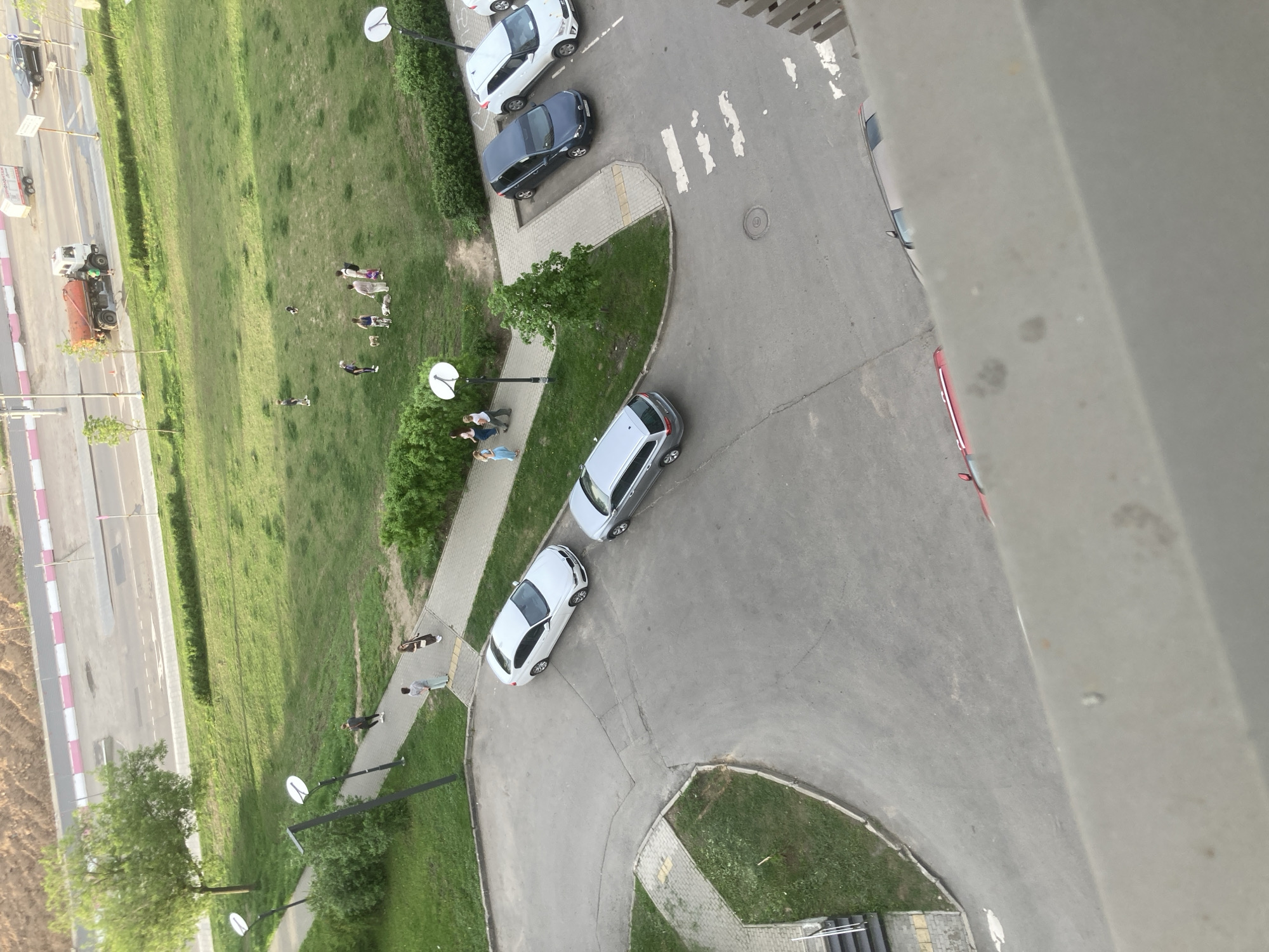 An aerial view shows a residential area in Khimki, Russia. A grassy field occupies the left side of the image, bordered by a road. On the grass, several people are visible, some standing and others walking. Adjacent to the grass is a narrow paved path. Cars are parked on the road and in parking areas. There are satellite dishes on poles in the grassy area. A large building is partially visible on the right side of the image.