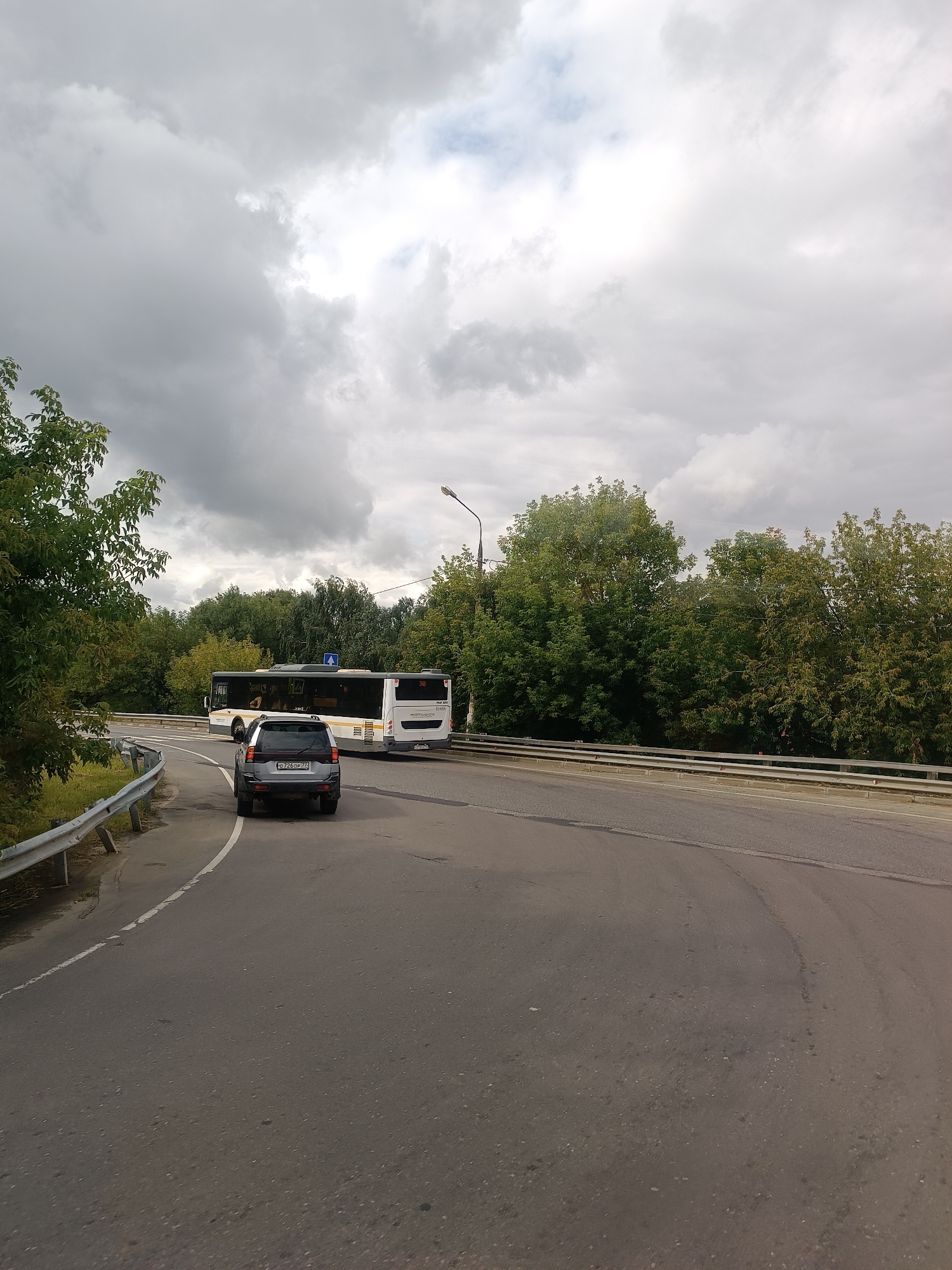 In Tokaryovo, Russia, a grey SUV is observed from behind on a paved road that curves to the right. Behind the SUV, a white bus with a dark stripe along its side is positioned on the same road, facing left. Both vehicles are under a cloudy sky. Lush green trees form a dense backdrop on either side of the road. A metal guardrail runs along the left edge of the road.