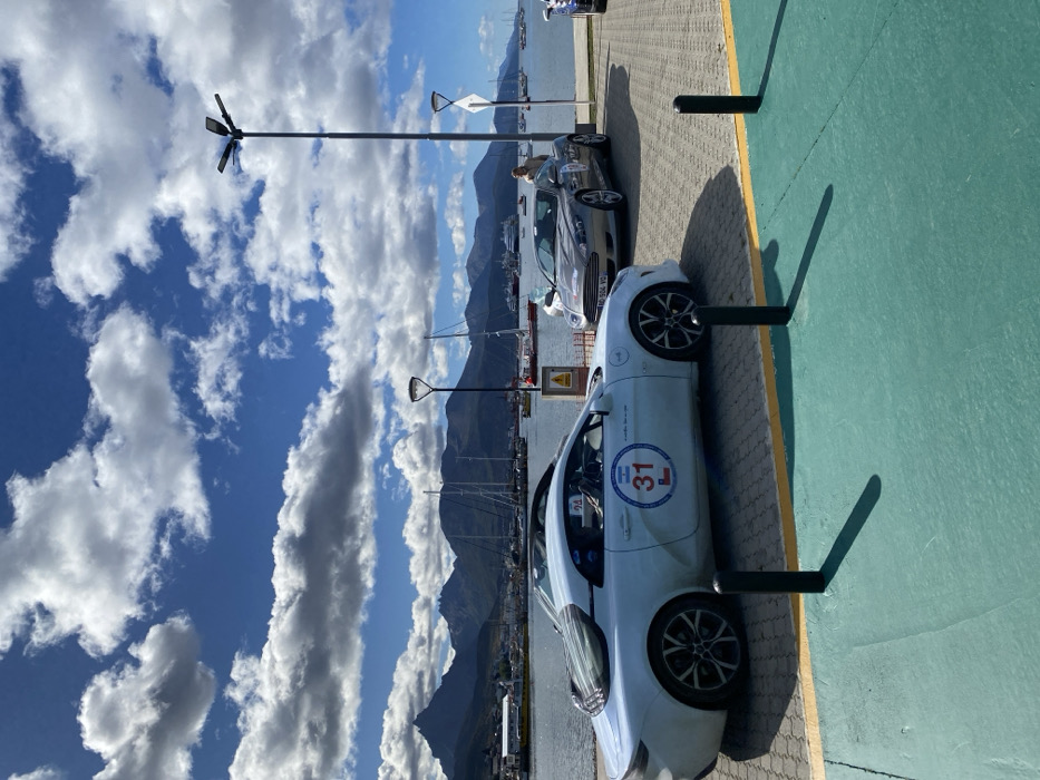 The scene depicts a waterfront area in Ushuaia, Argentina, under a bright blue sky with scattered white cumulus clouds. In the immediate foreground, a teal-green painted surface, likely a pedestrian or bike path, is separated by a thin yellow line from a grey paved area. Two black cylindrical bollards are positioned on the grey pavement.
Two luxury sports cars are prominently featured:
1. A white Alpine A110 sports coupe is parked on the left, facing towards the left. It bears a large circular decal on its door displaying the number "31," an Argentine flag, and the letter "L." The car has multi-spoke dark alloy wheels and a louvre-style rear window.
2. A silver Aston Martin DB9 Volante convertible is parked to the right and slightly behind the Alpine. Its French license plate "AD-504-YG" is visible. A less distinct circular decal is also present on its door.
One person, dressed in light-colored clothing, stands next to the driver's side of the silver Aston Martin, observing the vehicle.
Between the two cars, a yellow triangular warning sign with a black exclamation mark is visible. Several modern streetlights are positioned throughout the scene.
The midground features a calm body of water, likely a bay or harbor, populated with numerous moored sailboats and a larger, multi-decked vessel in the distance. Low-rise waterfront buildings are partially visible along the shore. The background is dominated by a range of dark, jagged mountains characteristic of the Patagonian landscape.