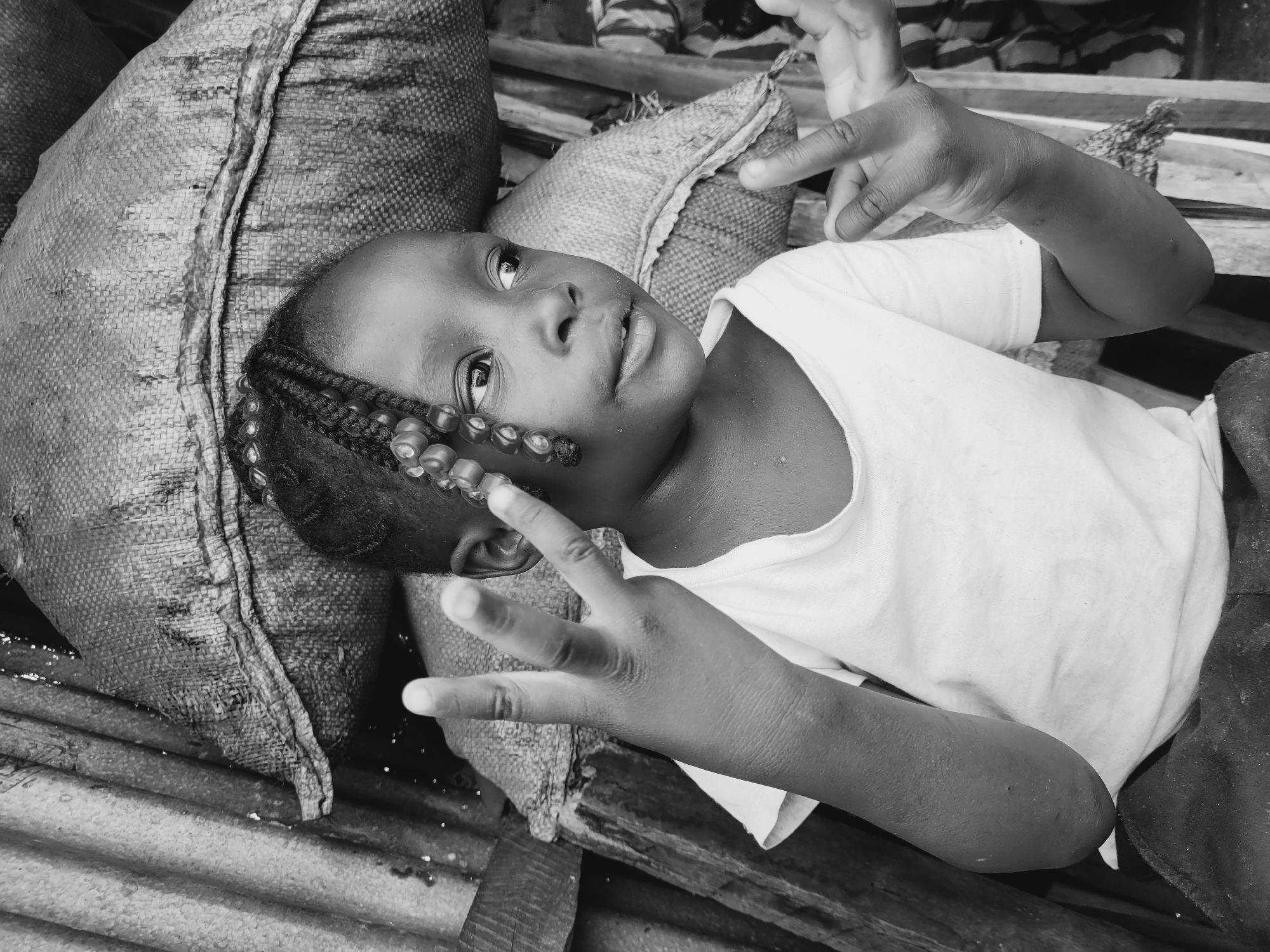 A black and white photograph captures a young child lying on a corrugated metal surface, presumably on the ground, in Bukuru, Nigeria. The child is positioned on what appears to be a burlap sack, which is used as a makeshift pillow. The child has braids with beads and is wearing a light-colored shirt. The child is gazing upward while holding their hands up, fingers spread apart, in a gesturing manner. The backdrop consists of what appears to be wood and various objects.
