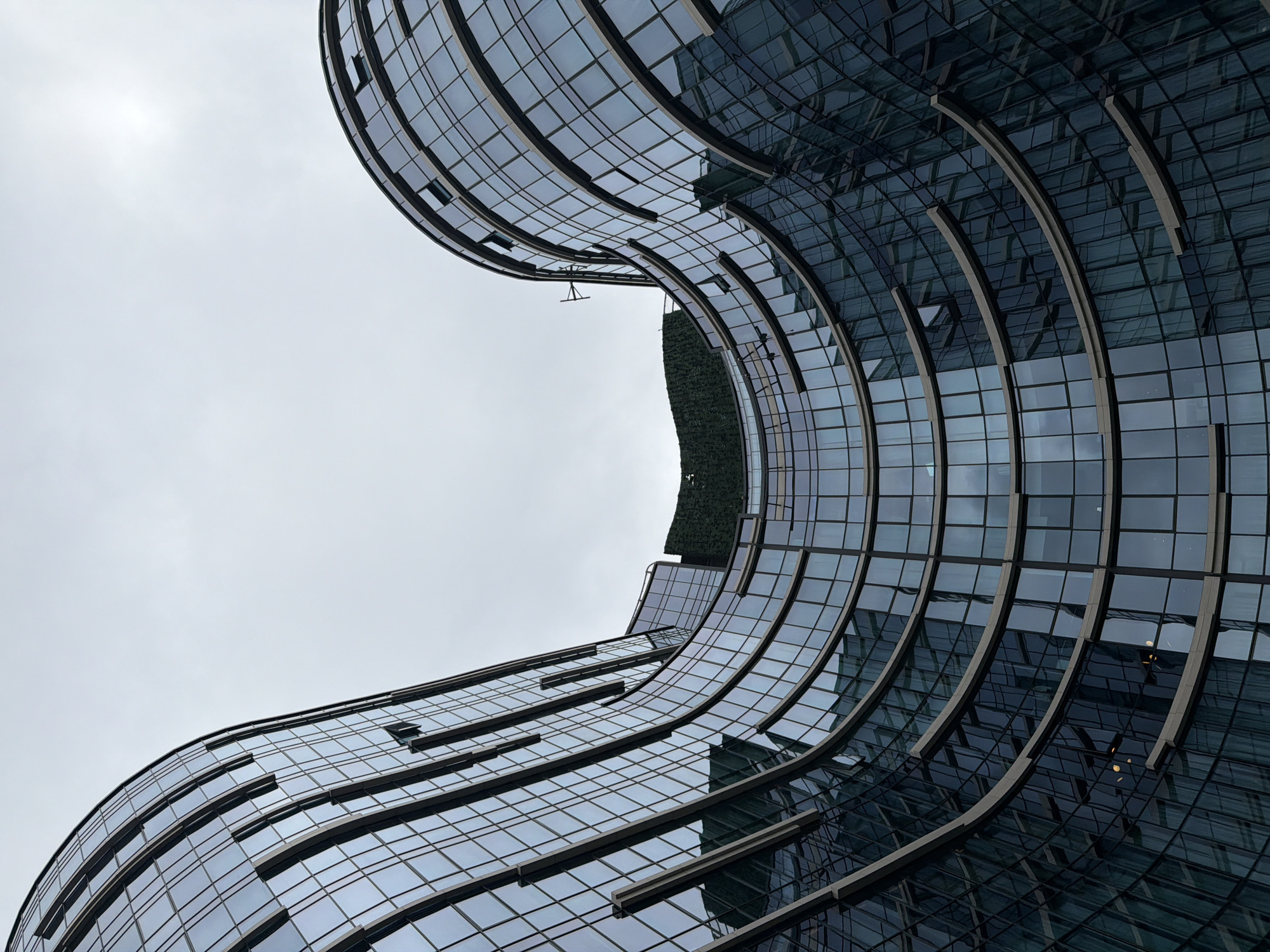 The image depicts the undulating, glass-facade exterior of a modern building in Tbilisi, Georgia. The architecture features curved, layered sections of reflective windows, creating a sense of movement and depth. The sky above is overcast and a pale gray, with no visible sun. A strip of green foliage is visible at the top of the structure, suggesting a rooftop garden or vertical landscaping. The perspective is from below, looking up at the building, emphasizing its grand scale and organic form. No people or vehicles are present in the frame. The building's design appears to be a contemporary interpretation of architectural elements, possibly designed to maximize natural light or create a unique aesthetic within the urban landscape.