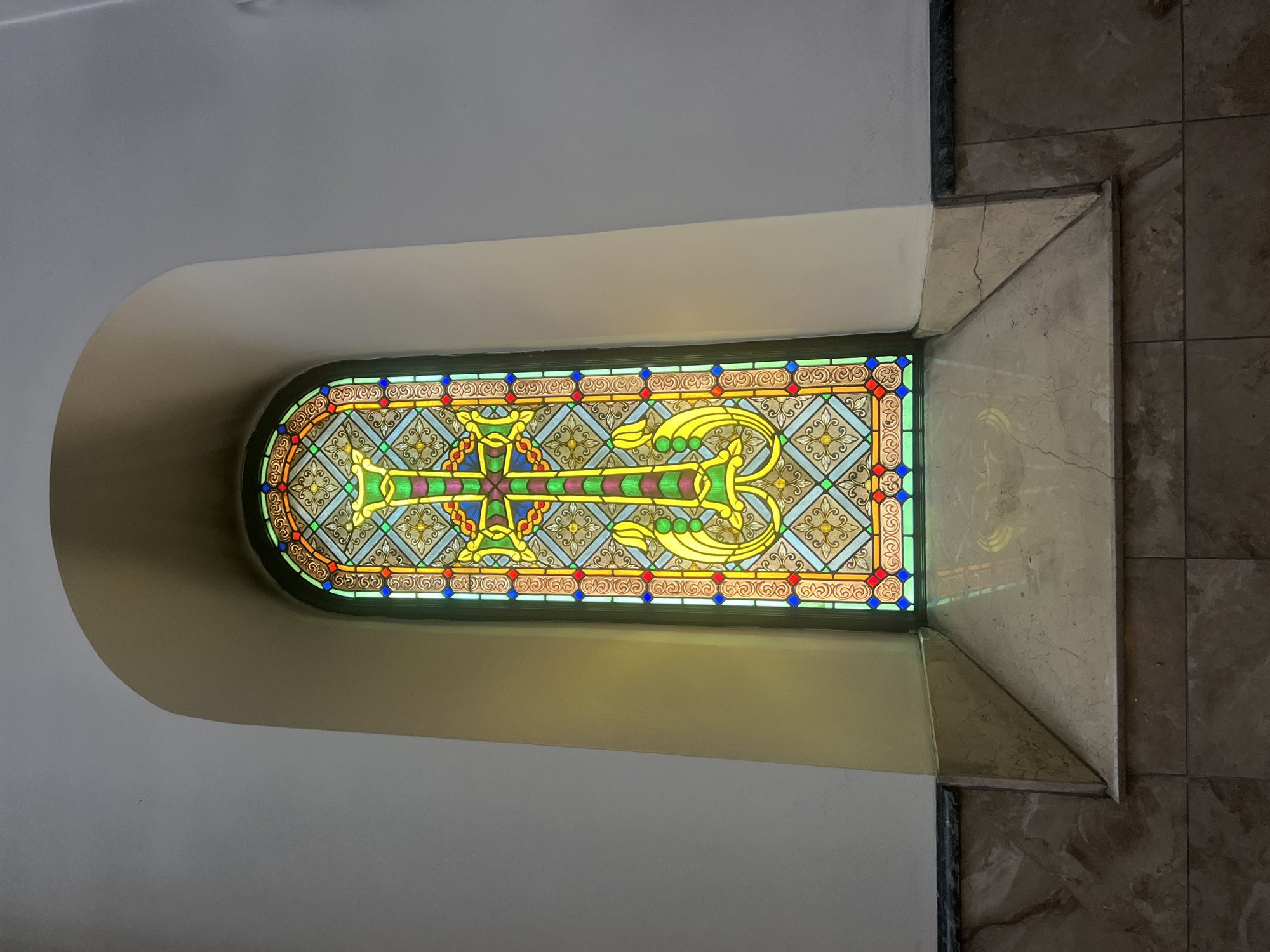A stained glass window featuring a prominent cross design is set into an arched alcove. The window is tall and narrow, with vibrant colors including green, yellow, red, and blue. The cross itself is primarily green and yellow, with intricate patterns surrounding it. Below the window, a marble windowsill forms a triangular shelf. The wall surrounding the alcove is painted a light cream color. The lower portion of the wall is clad in marble and tile. The overall scene suggests an interior setting, likely a religious building.