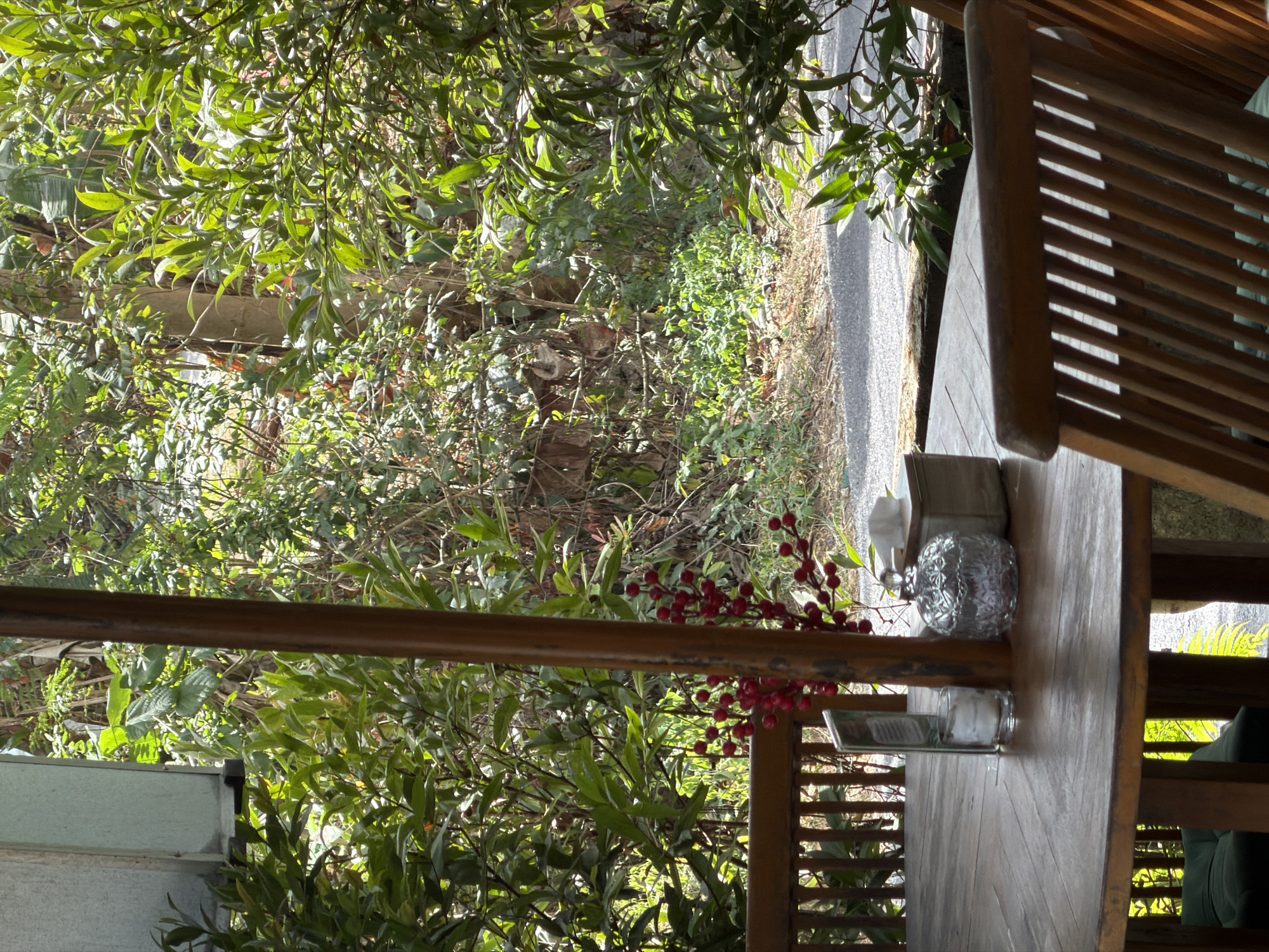 The image depicts a wooden table and chairs set in an outdoor dining area. The table is adorned with a small glass candle holder, a decorative glass container, a box of tissues, and a sprig of red berries. Behind the table, lush green foliage and trees form a natural backdrop, with a glimpse of a road or pathway visible in the distance. The scene suggests a tranquil, nature-immersed setting. The location is identified as Kuta Selatan, Indonesia.