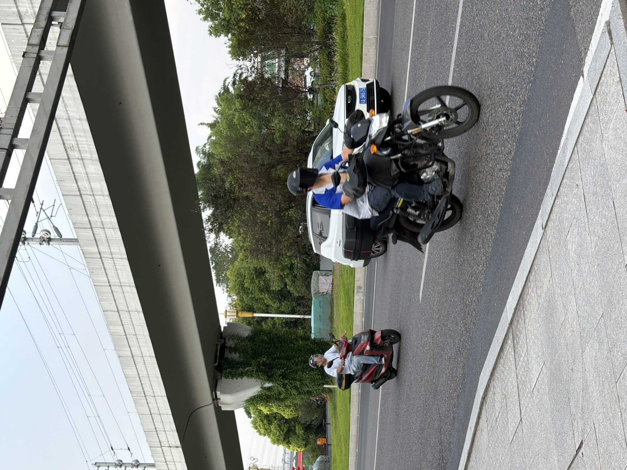 In Jiangjin, China, two individuals are riding motorcycles on a multi-lane paved road under an elevated structure. The person in the foreground, on a black motorcycle, wears a blue and white shirt, dark pants, and a full-face black helmet. They are riding in the right lane. Behind them and to the left, another person on a red scooter wears a white t-shirt, blue jeans, and a white helmet with a clear visor, also riding in the right lane. A white SUV is visible in the left lane, and a white line marks the lane divisions. Lush green trees and vegetation line the side of the road, with a concrete sidewalk in the foreground. Overhead, a concrete bridge or overpass supports visible electrical wires. The sky is overcast.
