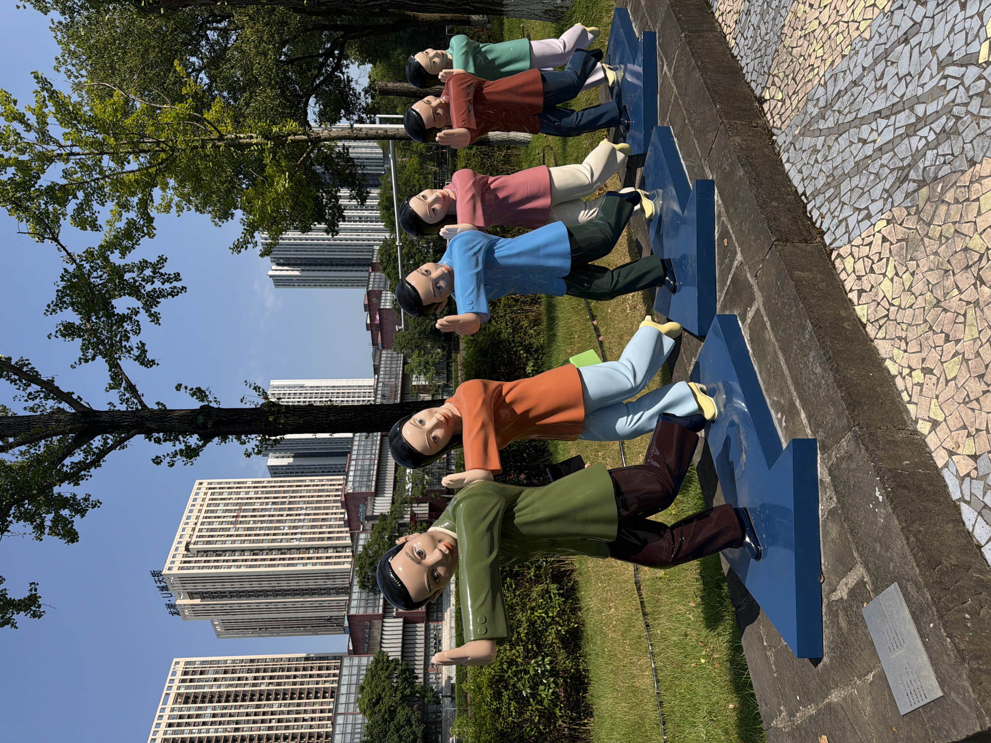 In a sunny outdoor environment, a series of six human-like statues stand in a line, each with a determined expression and arms extended forward in a pushing motion. The statues are arranged on blue arrow-shaped bases, arranged diagonally and receding into the background, implying movement or direction. The figures are dressed in colorful clothing: the first statue from the left wears a green jacket and brown pants, followed by an orange jacket and light blue pants, a blue jacket and dark green pants, a pink jacket and light beige pants, a maroon jacket and dark blue pants, and finally a light green jacket and light purple pants. The statues are positioned on a paved walkway with a mosaic pattern of light-colored tiles. Behind them, a grassy area is bordered by a low stone wall. In the background, several modern high-rise buildings, some with prominent signage, are visible under a clear blue sky, suggesting an urban setting. The overall scene depicts a dynamic representation of collective effort or progress, set against a backdrop of contemporary architecture. This installation is located in the Chongqing High-tech Zone, China.