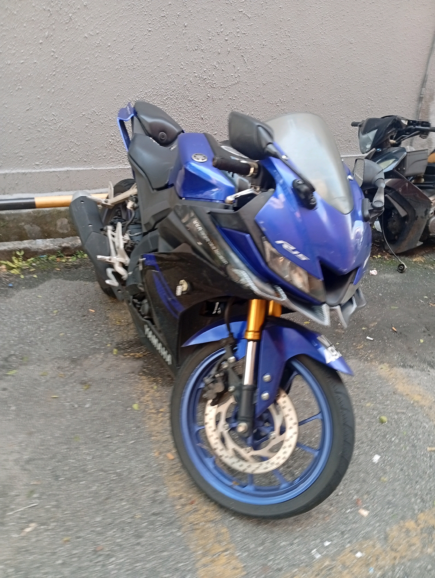 A blue Yamaha R15 motorcycle is parked on an asphalt surface next to a grey textured wall. The motorcycle is angled slightly towards the viewer and is the primary subject of the image. The front wheel is clearly visible with its blue rim and silver brake disc. Gold-colored front suspension forks are also prominent. The motorcycle's bodywork is a vibrant blue, with black accents. A partially obscured black motorcycle is visible behind and to the right of the Yamaha. The background consists of a grey, textured wall with a yellow and black striped barrier running along its base. The ground is a grey asphalt surface with scattered leaves and debris. There are no people visible in the image. The scene appears to be an outdoor setting in an urban or semi-urban environment, consistent with a location in the Selayang Municipal Council, Malaysia.