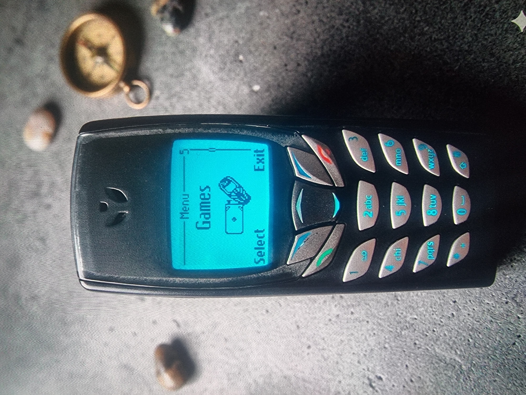The image depicts a Nokia mobile phone lying on a textured gray surface. The phone's screen is illuminated with a blue backlight, displaying the word "Games" under the "Menu" option. The screen also shows prompts for "Select" and "Exit". The phone's keypad has individual buttons with alphanumeric labels, such as "1", "2 abc", "3 def", etc., with the numbers and letters highlighted in blue. To the left of the phone, in the background, are a small compass and a few small, irregularly shaped items. The specific location or city is not discernible from the image.
