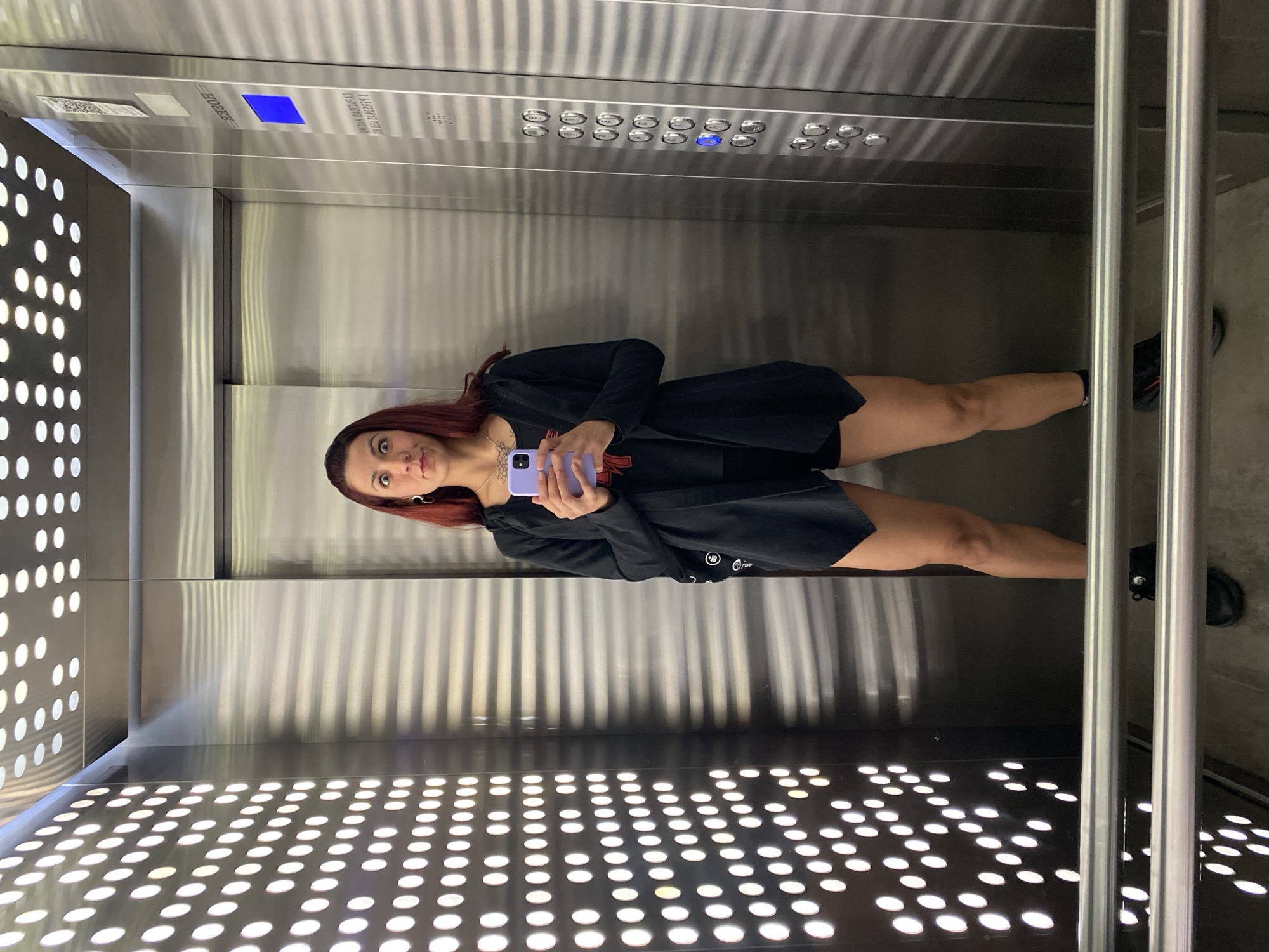 The scene depicts a solo female subject taking a mirror selfie inside a modern elevator located at Av. Ing. Huergo 1121, Puerto Madero, Buenos Aires, Argentina. The elevator features brushed stainless steel walls, with a horizontally ridged pattern on the back and right walls. The left wall and ceiling are adorned with evenly spaced, illuminated white circular lights. A metallic handrail with two parallel bars is present in the lower foreground. The female, appearing to be in her mid-20s to mid-30s, has long, dark red hair, silver hoop earrings, and a visible tattoo on her upper chest. She is dressed in a black ensemble consisting of shorts, a t-shirt (with a red detail partially visible), an open, long-sleeved black cardigan with an asymmetrical hem, and dark athletic shoes with red accents. She holds a light purple smartphone at chest level, directing it towards her reflection. Her facial expression is exaggerated, characterized by wide eyes, raised eyebrows, and pursed lips. The elevator control panel, visible on the right wall, displays a blue floor indicator, a vertical column of circular buttons (one illuminated blue), a QR code, and mirrored text indicating "HOFER" and the address "AV. ING. HUERGO 1121 BUENOS AIRES".