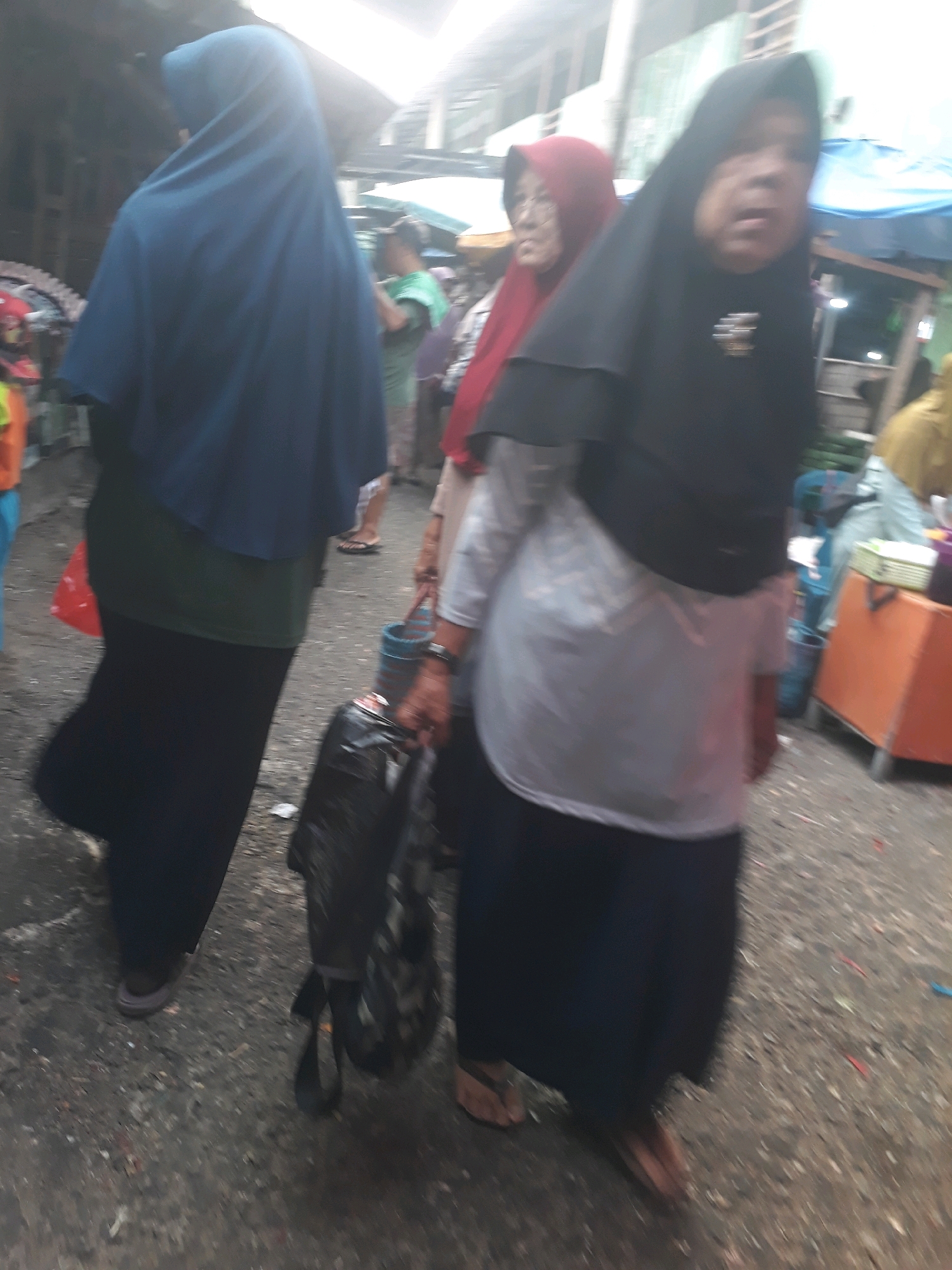 In Pangkalpinang, Indonesia, three individuals are depicted in a market setting. The foremost person, on the right, wears a black hijab and a white blouse, carrying a black backpack. To their left, another person in a red hijab is visible, their facial features slightly blurred. The third person, on the far left, is seen from behind, wearing a dark blue hijab and a dark skirt. The ground appears to be paved with dirt and debris, indicative of an outdoor market. In the background, a blue tarp covers a stall, and a wooden cart with orange drawers is present, suggesting commercial activity. The overall scene is characterized by motion blur, indicating movement within the market.