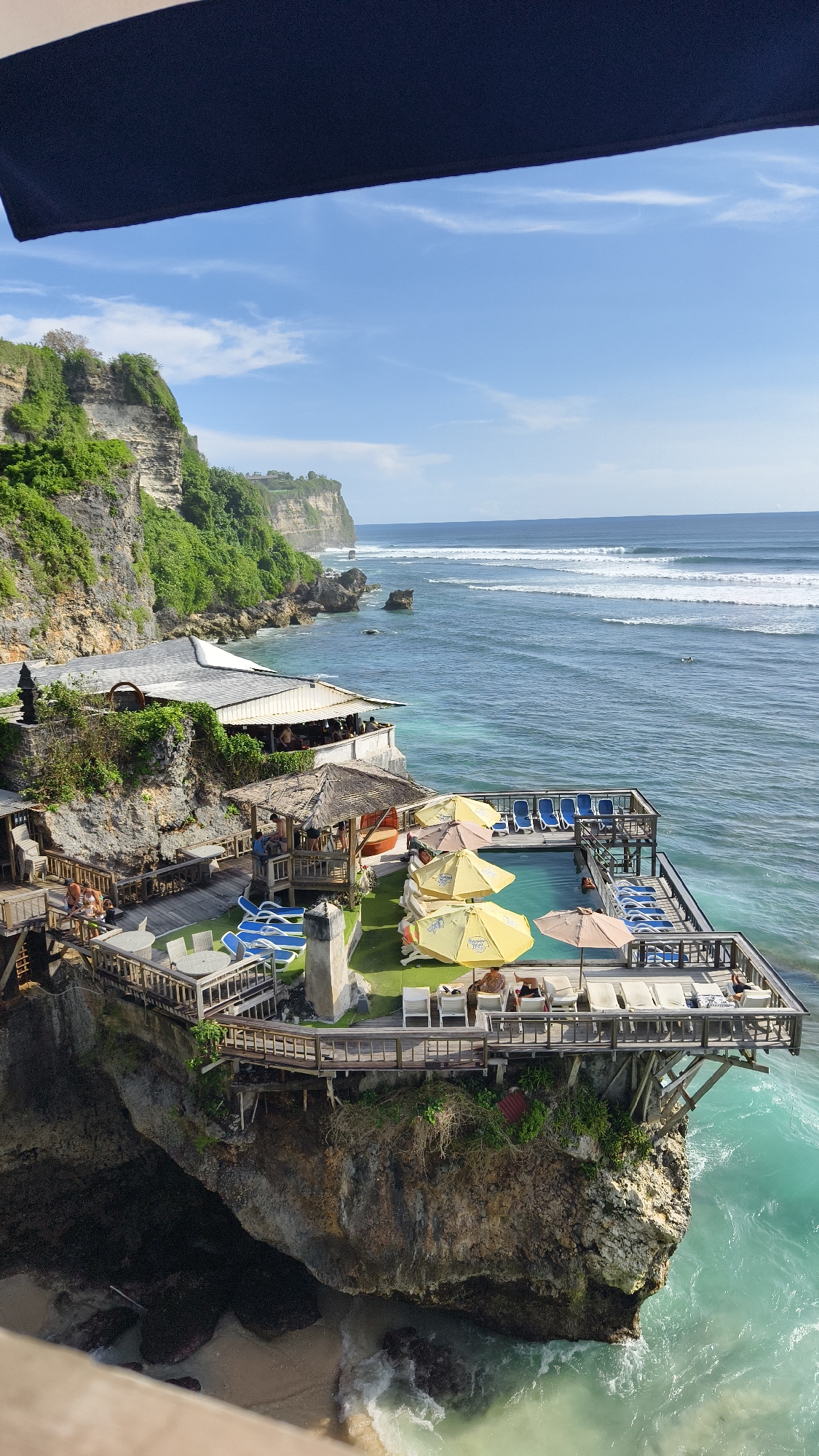 The image captures a high-angle view of Uluwatu, a coastal area in Pecatu, Indonesia. The scene features a multi-tiered cliffside complex with a restaurant and a swimming pool deck. The restaurant is situated on the edge of the cliff, with its white roof visible. Below it, there is a wooden deck area with tables and seating. Adjacent to this, there is a swimming pool surrounded by a deck area equipped with lounge chairs and yellow umbrellas. People are visible on the deck, either swimming or relaxing on the lounge chairs. The pool deck extends out over the cliff edge, offering views of the ocean. The ocean is a gradient of blue hues with visible waves. The sky is blue with light clouds. The cliff is composed of layered rock formations with green vegetation growing on top and along its sides. The foreground reveals a part of another structure.