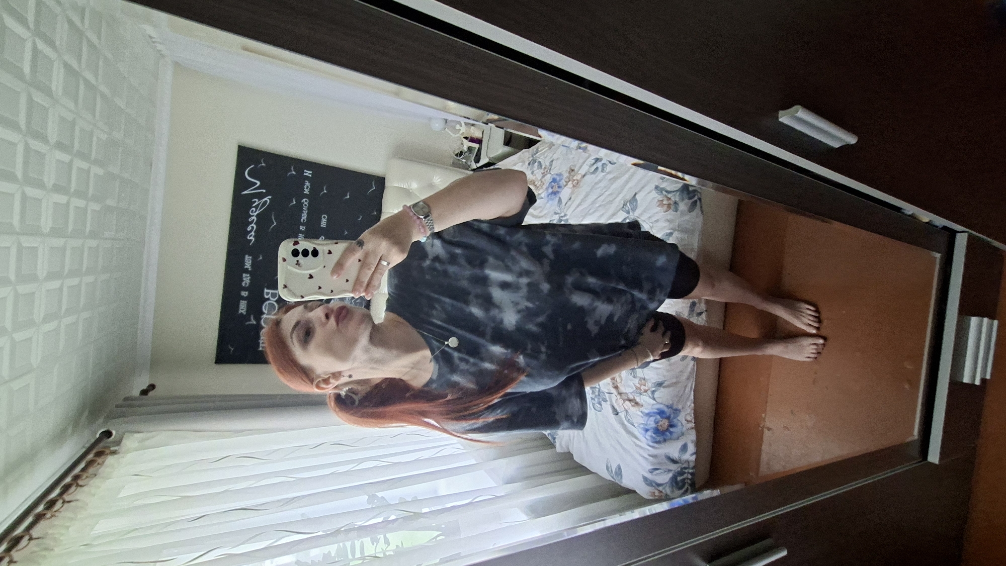 A woman with long, auburn hair pulled into a ponytail is reflected in a mirror. She is wearing a dark tie-dye t-shirt and dark shorts. She is holding a smartphone in her right hand and making a kissing face. The background of the reflection shows a bed with a floral comforter and a window with sheer curtains. A dark framed wardrobe is visible to the right of the mirror. The image was taken in an indoor setting, possibly a bedroom, and does not provide any specific details about the city of Myadzyel, Belarus.