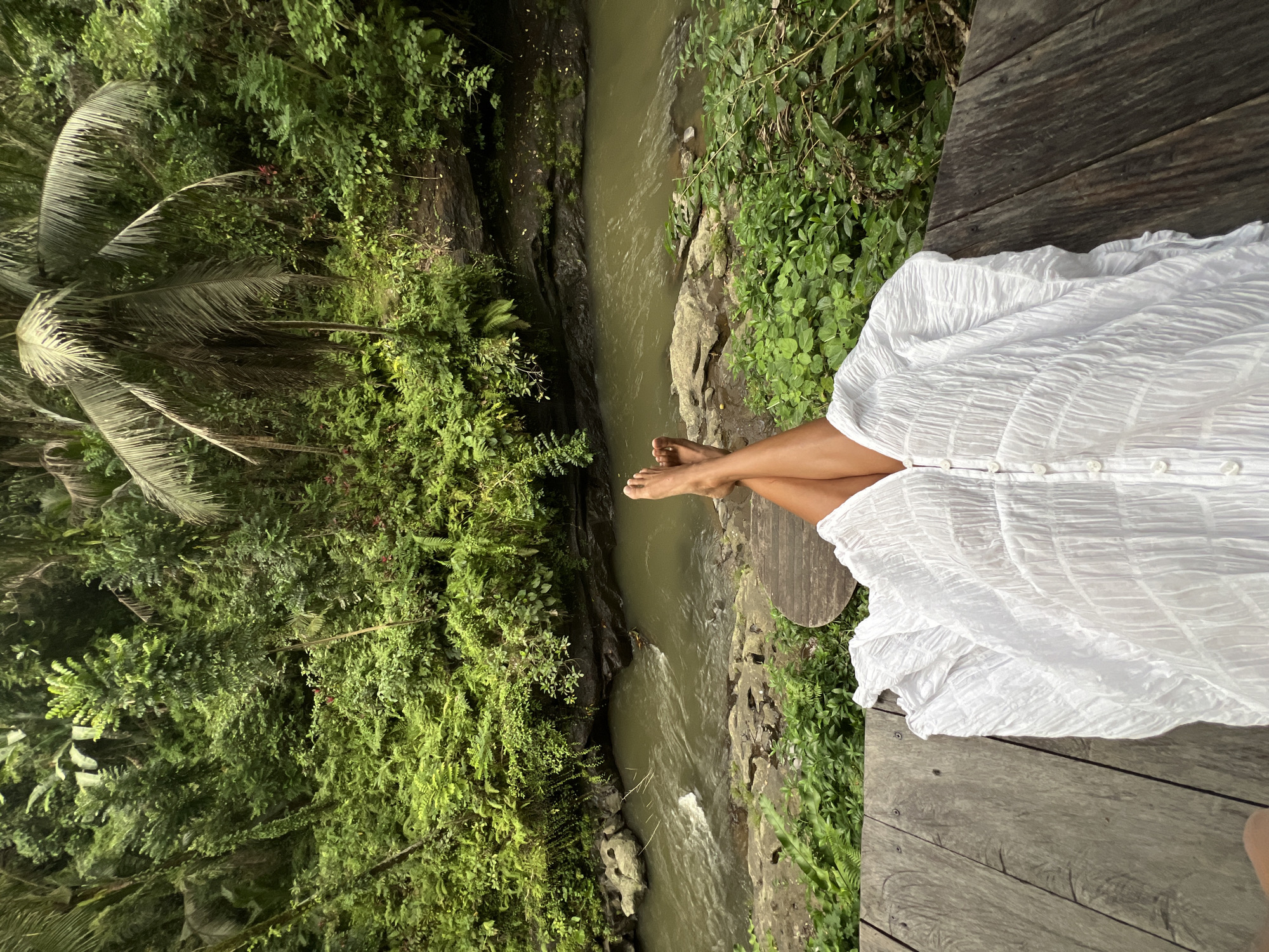 The image depicts a person sitting on a wooden platform overlooking a lush green jungle and a river flowing through it. The person is wearing a flowing white dress and their legs are crossed, with their bare feet visible. The setting appears to be a tropical rainforest, with dense vegetation, palm trees, and ferns surrounding the river. The river itself is murky green and appears to be flowing rapidly in some areas. The wooden platform is made of weathered planks, suggesting it is an outdoor structure designed to provide a vantage point for enjoying the natural scenery.
The lighting suggests it is daytime, likely in the late afternoon or early morning, given the dappled sunlight and shadows. The overall mood is tranquil and serene, evoking a sense of peace and connection with nature. The location, identified as Ubud District, Indonesia, further reinforces the tropical jungle setting. There is no visible text in the image. The focus is on the natural beauty of the landscape and the relaxed posture of the person enjoying it.
