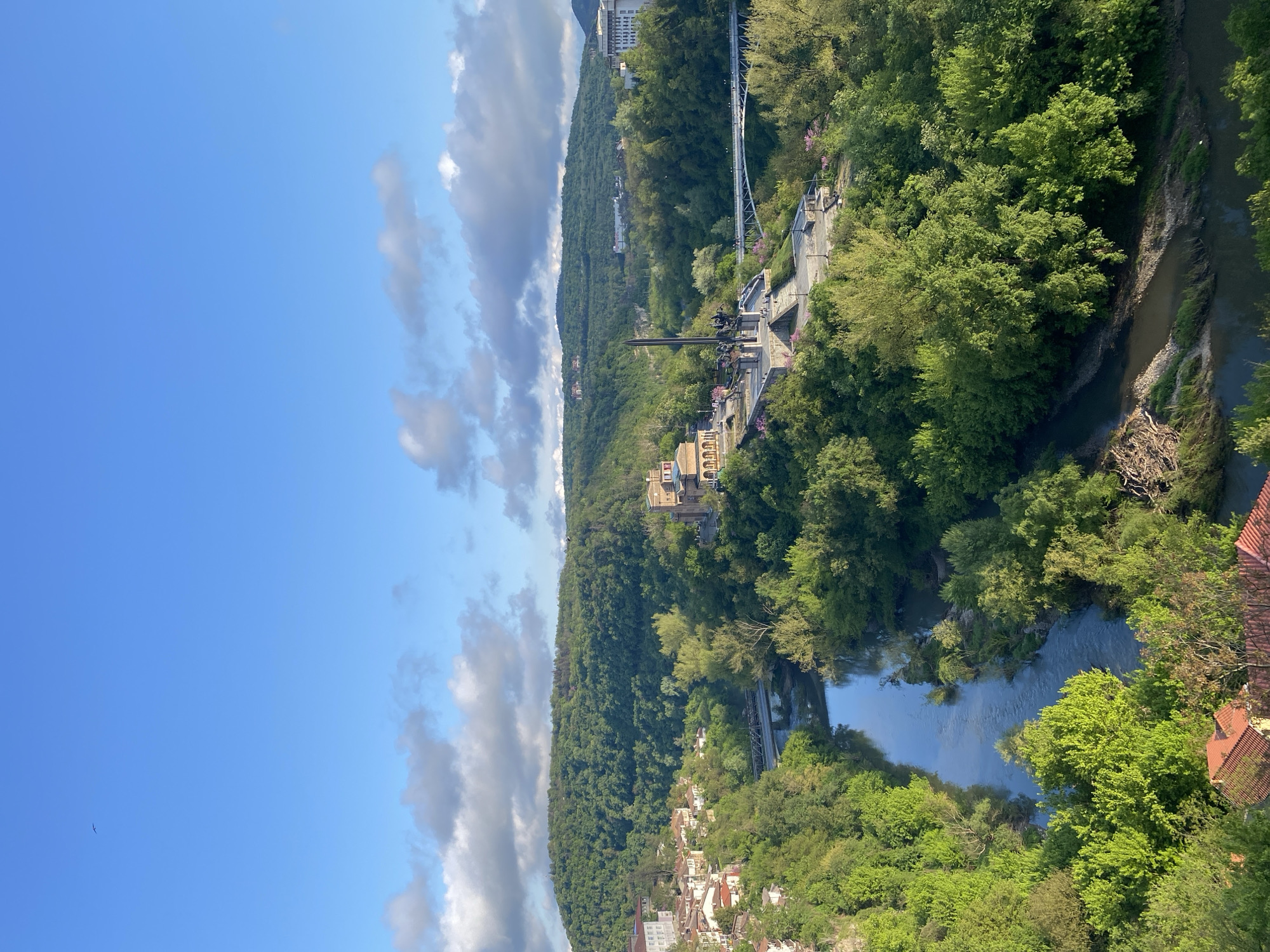The image presents a landscape featuring a river winding through a lush, green valley. On one side of the valley, a series of buildings are situated, some of which appear to be historical structures. A pedestrian bridge spans across the valley connecting the two sides. The sky above is a mix of blue and white clouds. The location appears to be in the city of Veliko Tarnovo in Bulgaria.