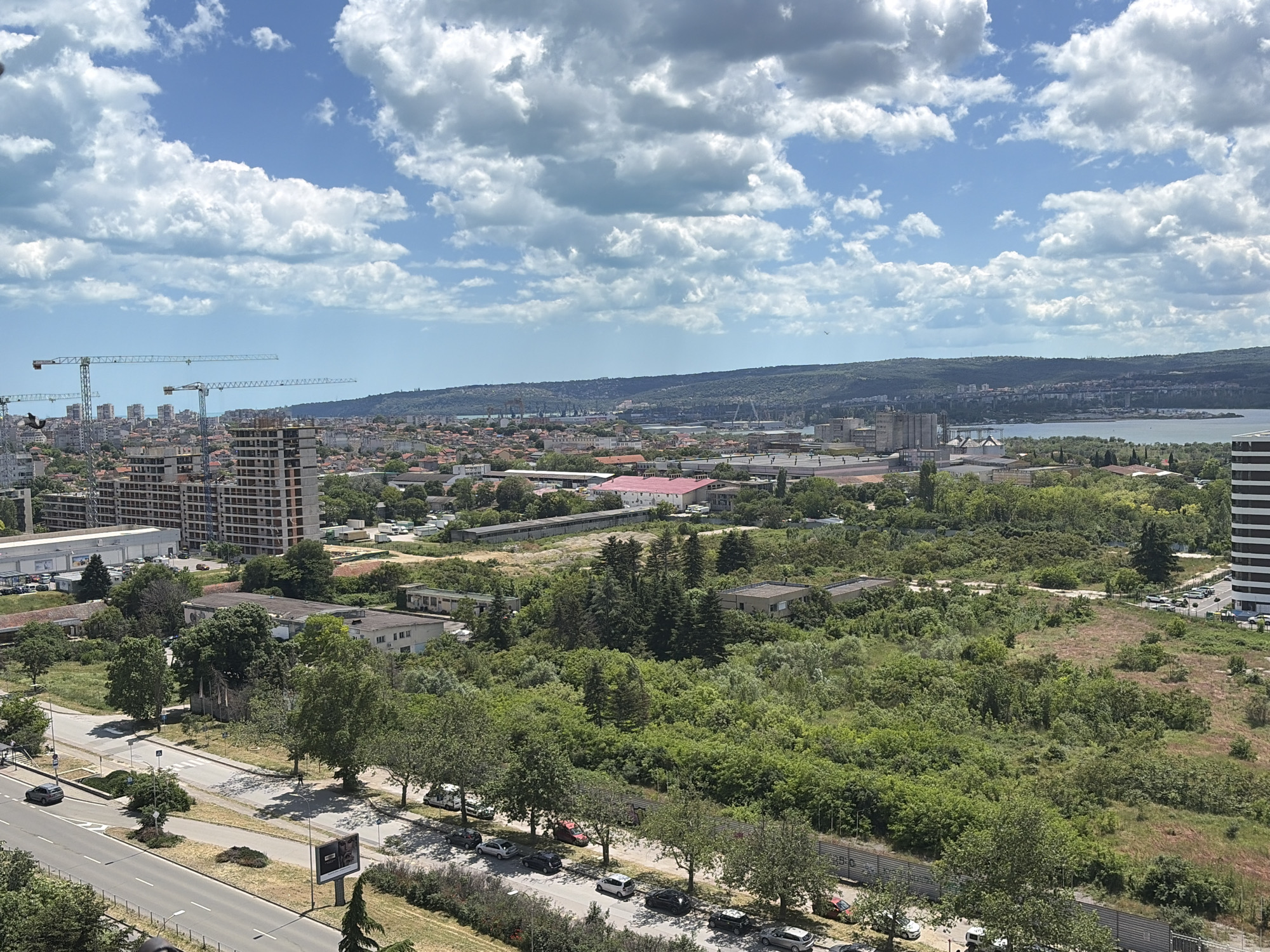 The image captures a daytime view of Varna, Bulgaria, under a partly cloudy sky. In the foreground, there's a multi-lane road lined with trees and parked cars. Behind the road, dense greenery transitions into a cityscape with buildings of varying heights, including a tall striped building on the right edge and buildings with cranes in the distance. The terrain rises gradually toward the background, where a hill and the coastline meet the sea. The overall impression is a mix of urban and natural landscapes under a bright, summer-like sky.