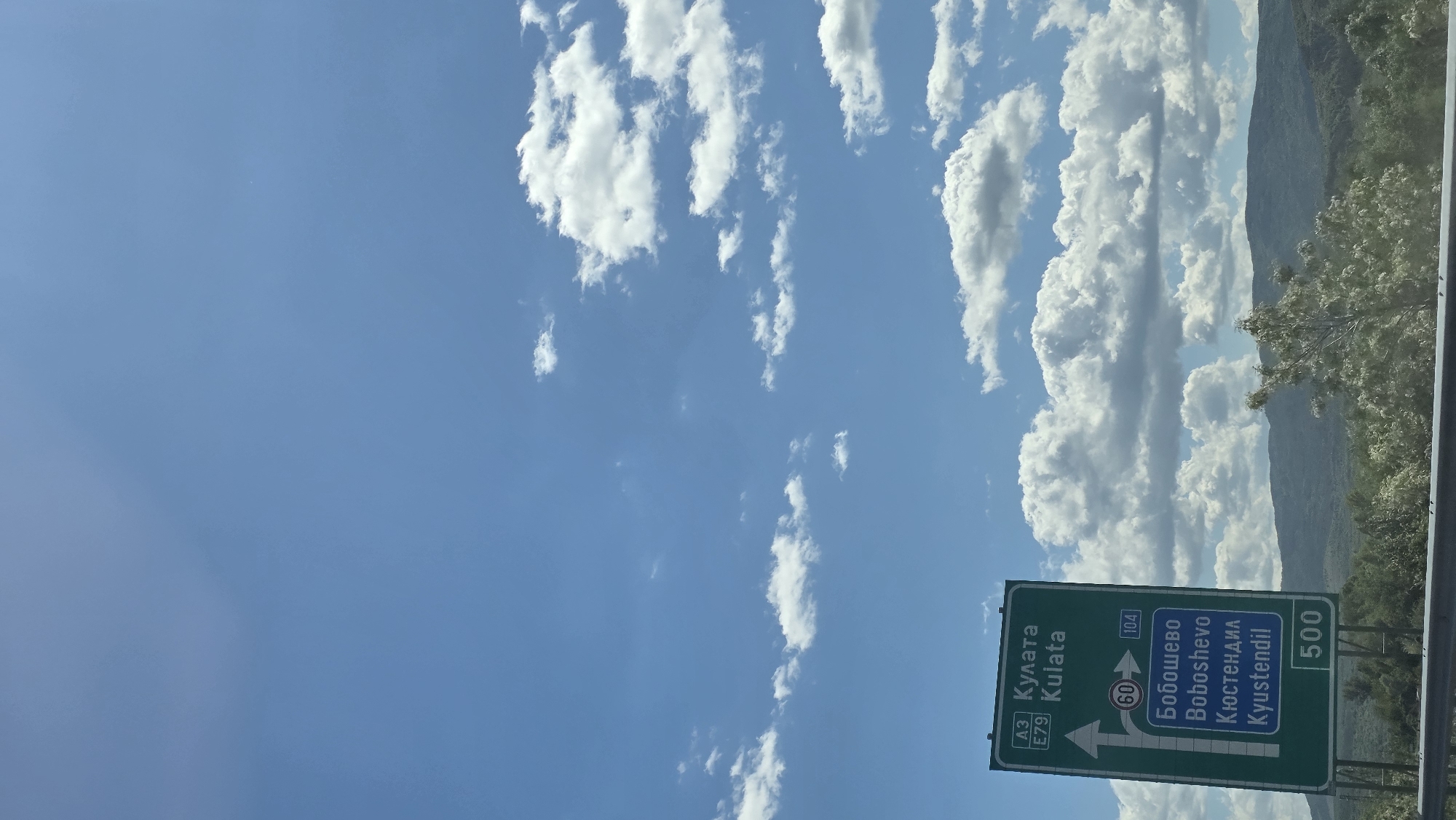The image depicts a daytime scene on a highway in Bulgaria, near the city of Blajievo. Dominating the view is a blue sky with scattered white cumulus clouds. A green highway sign is visible, indicating directions to: "Кулата Kulata" (left), "Бобошево Boboshevo" (straight), and "Кюстендил Kyustendil" (straight). The highway numbers are A3 and E79 for Kulata, and 104 for Boboshevo and Kyustendil. A speed limit sign of 60 is visible above the directional arrow. The sign also indicates that Kyustendil is 500 kilometers away. In the background, there is a mountain range with vegetation. The image appears to be taken from inside a vehicle.