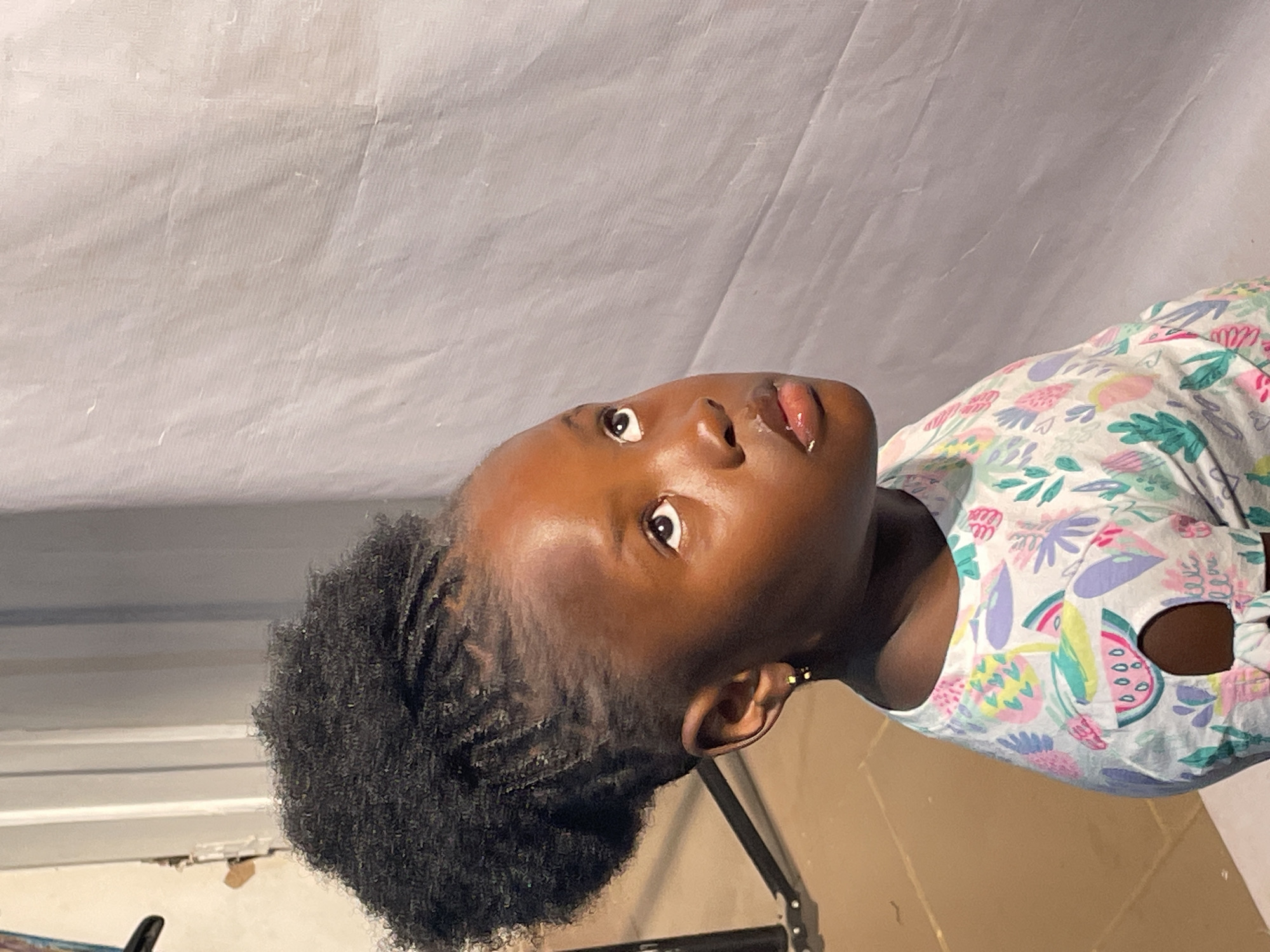A young Black girl with dark, curly hair pulled up in a high ponytail is shown from the chest up, looking upwards and to the right. She has a small gold hoop earring in her left ear. She is wearing a white t-shirt with a colorful pattern of green leaves, pink and purple flowers, and slices of watermelon with seeds. Her lips are glossy. The background is a textured, light gray fabric. To the left of the girl, a black tripod is visible. The scene appears to be indoors, possibly a studio setting. The context of Jos, Nigeria, is not discernible from the visual information provided in the image.