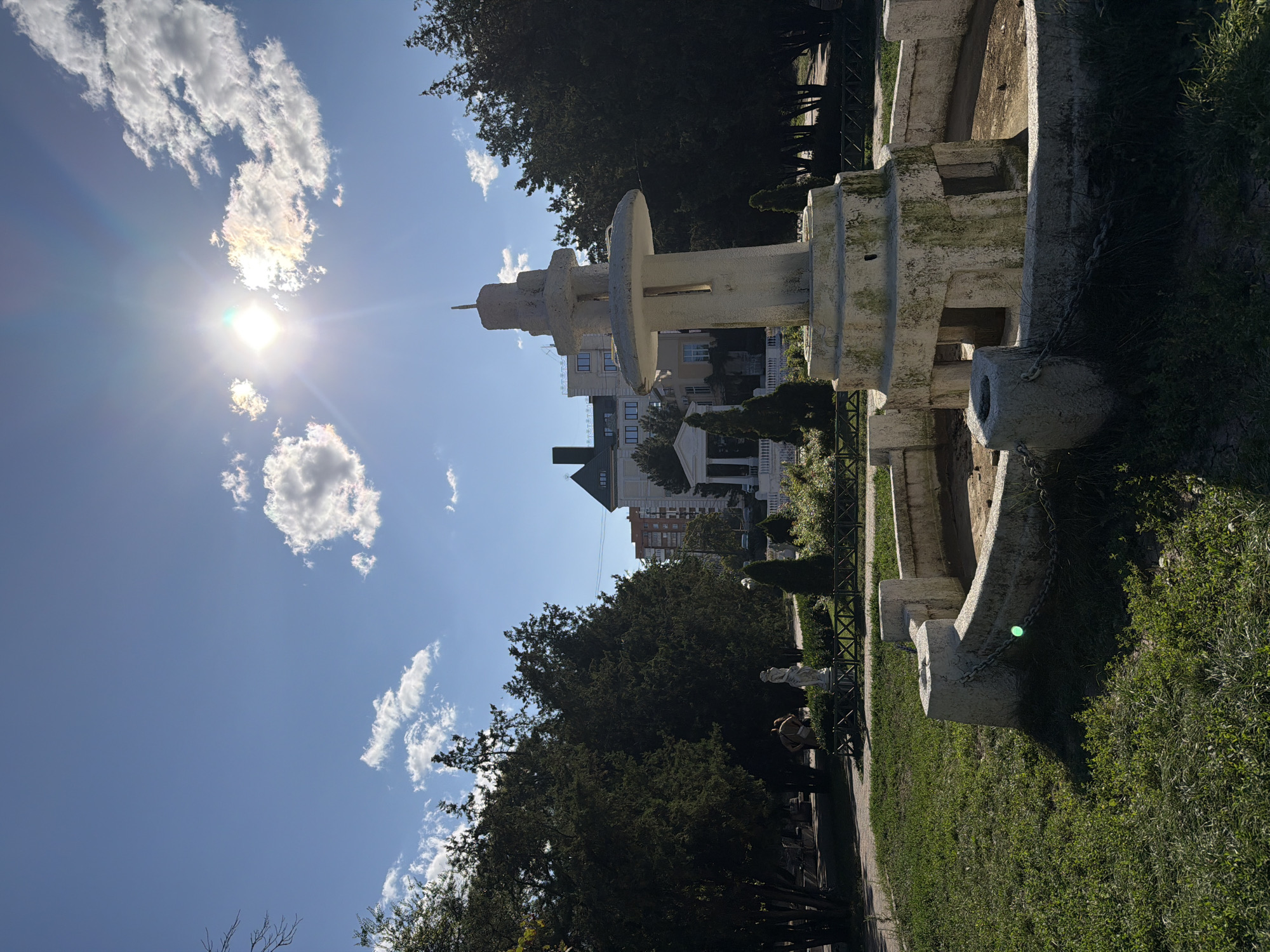 The image depicts an outdoor scene in a park setting in Rostov-on-Don, Russia, likely during daylight hours due to the bright sunlight and clear sky with some scattered clouds. In the foreground, there is an old, dry fountain with a weathered, multi-tiered stone structure at its center. The fountain is surrounded by a low, stone border with chains draped around it. Green grass covers the ground around the fountain, with some patches of drier vegetation.
In the middle ground, there are several mature evergreen trees forming a dense border. Behind the trees, a white-columned structure, possibly a pavilion or part of a building, is visible. Beyond this, a multi-story building with a dark, pitched roof and windows can be seen. To the left of the central fountain, partially obscured by trees, is a white statue of a figure, and nearby, two individuals are standing, one appearing to be holding a bag. A wooden fence runs along the edge of a landscaped area.
The sun is high in the sky, casting a strong glare and creating lens flare. The overall impression is that of a public park, possibly historical, with elements of classical architecture.
