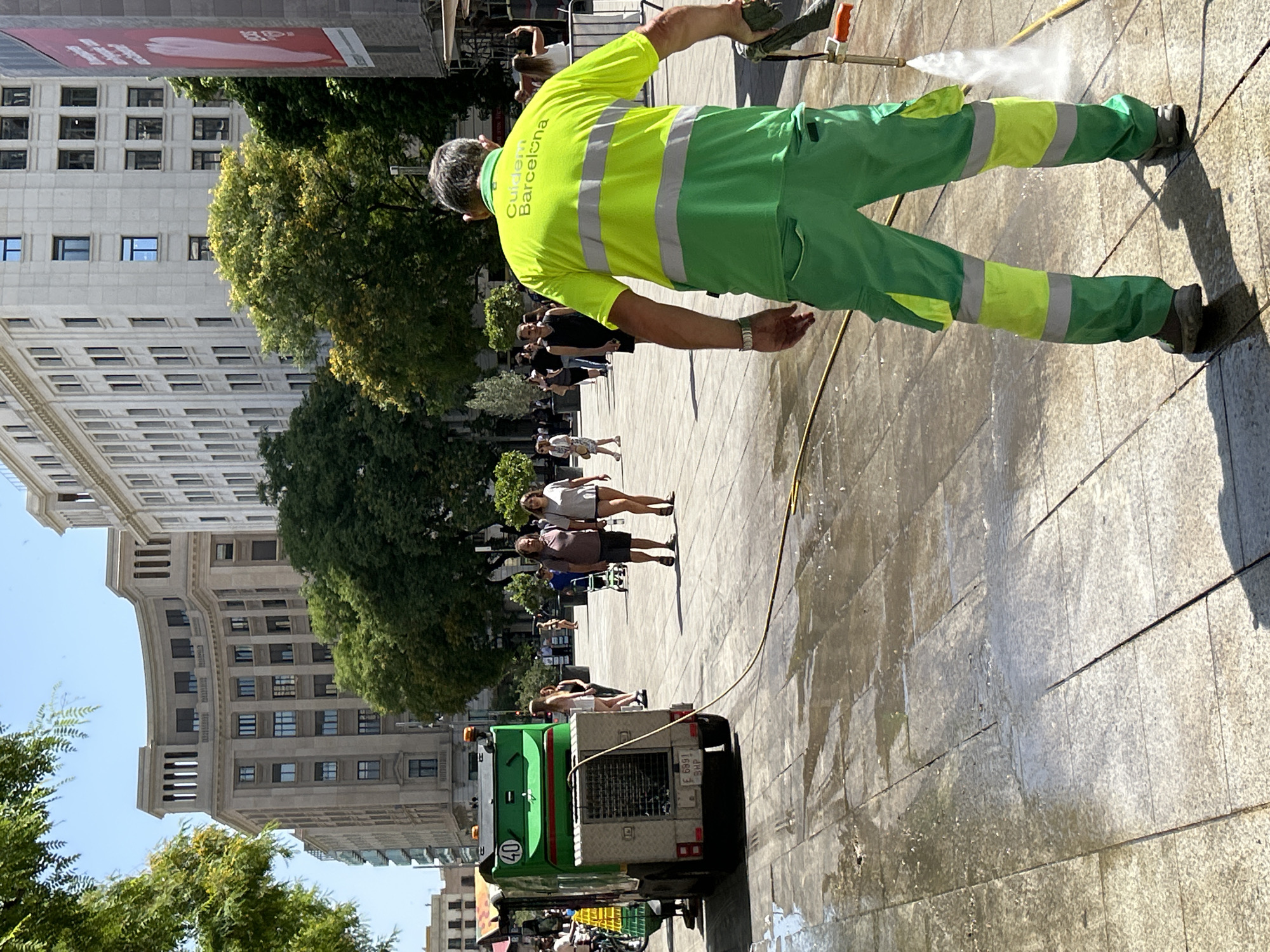 In Barcelona, Spain, a street cleaner wearing a bright green uniform with reflective stripes is pressure washing a tiled plaza. The cleaner is holding a spray wand attached to a hose, directing a stream of water onto the ground. To the left, a green cleaning vehicle with the number '40' displayed on its side is parked. In the background, several pedestrians are walking through the plaza, and large, ornate buildings line the street. Trees with lush green foliage are scattered throughout the scene, and the sky is clear and blue.