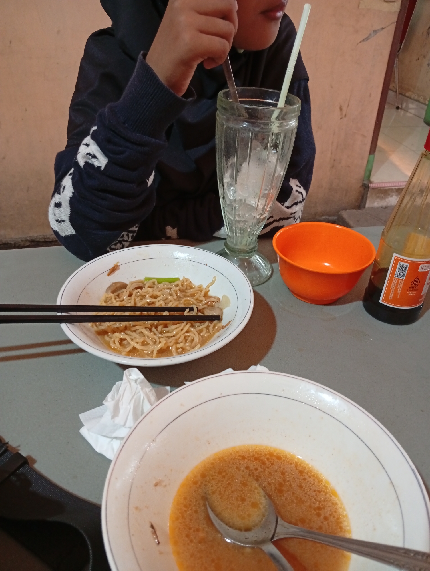 A person wearing a dark blue hoodie with white detailing on the sleeves is seated at a table, holding a spoon and a straw in a tall glass filled with ice. In front of them are two bowls of noodles, one containing a savory broth with meat and noodles, and the other appearing to be mostly broth with a spoon. An orange bowl sits to the right of the noodle bowls, and a bottle of soy sauce is also on the table. The scene is lit by artificial light, casting shadows. This image was taken in a restaurant in Curug Wetan, Indonesia.