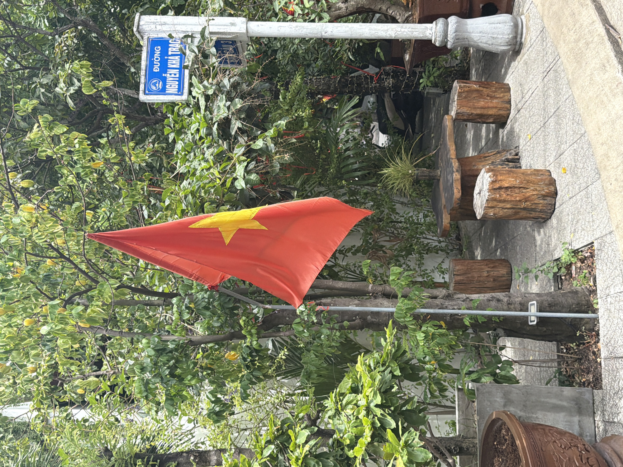 A Vietnamese flag, predominantly red with a yellow star, hangs from a pole in front of a lush green environment. The flag is positioned to the left of the frame. To the right of the flag, a white lamppost with a blue street sign is visible. The street sign displays Vietnamese text, reading "ĐƯỜNG NGUYỄN KHÁ TRẠC". This indicates the location is Nguyen Kha Trac Street in Hoi An, Vietnam. In the foreground, a small outdoor seating area is set up with a wooden table and three log stools. The ground is paved with grey tiles. Various potted plants and trees surround the scene, contributing to a natural and somewhat rustic ambiance. The overall impression is that of a street corner or a small public space in a Vietnamese city.