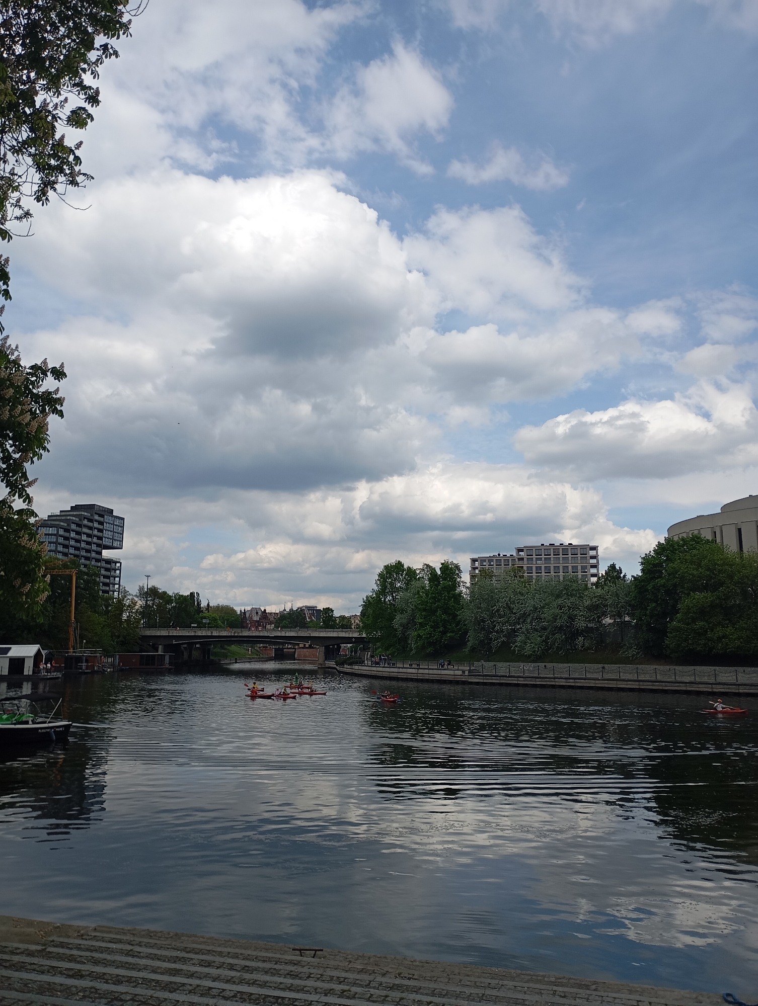 The image captures a waterway scene in Bydgoszcz, Poland. A body of water, likely the Brda River or a canal, stretches across the frame, reflecting the sky and surrounding structures. Several people are kayaking on the water. A bridge spans the waterway in the midground, connecting the two sides of the city. Buildings of varying heights and architectural styles line the waterfront, mixed with green foliage from trees. The sky is filled with clouds, indicating a partly cloudy day. The perspective is from a low vantage point, possibly from steps leading down to the water.
