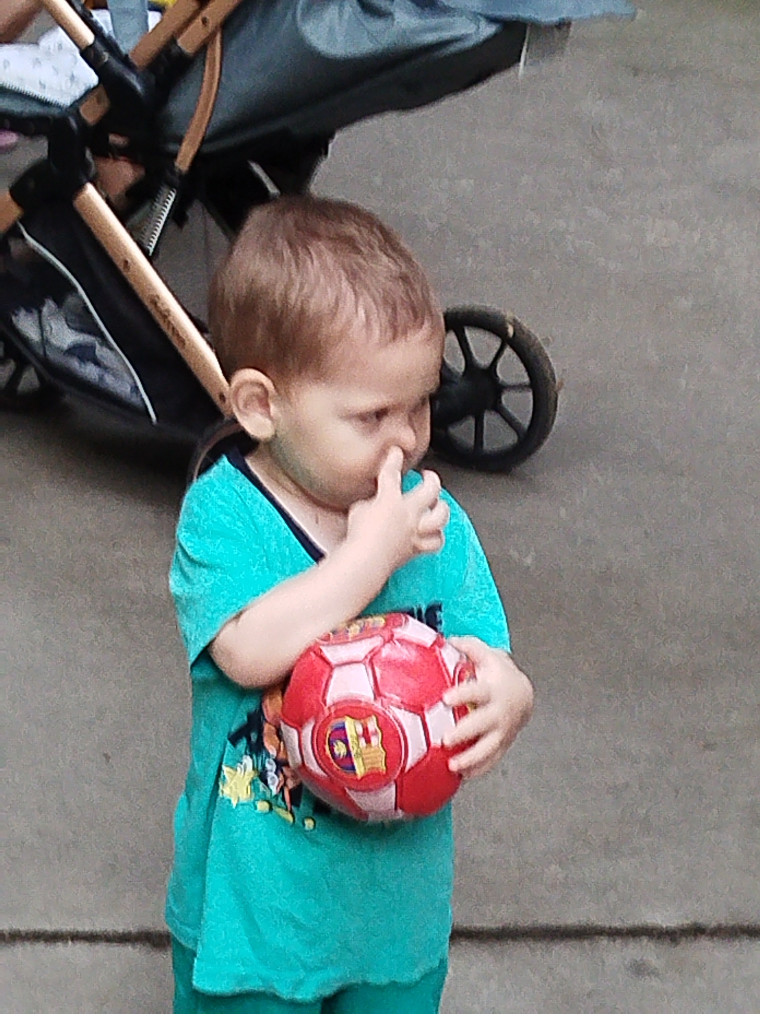 In the city of Unknown, Unknown, a young child, appearing to be male, stands outdoors and holds a red and white soccer ball. The child is wearing a teal t-shirt with a graphic design on the front and has light brown hair. The child is looking down and has their right hand to their nose. Behind the child, a stroller is visible, partially obscuring the background. The ground is a gray, paved surface. The lighting suggests it is daytime.