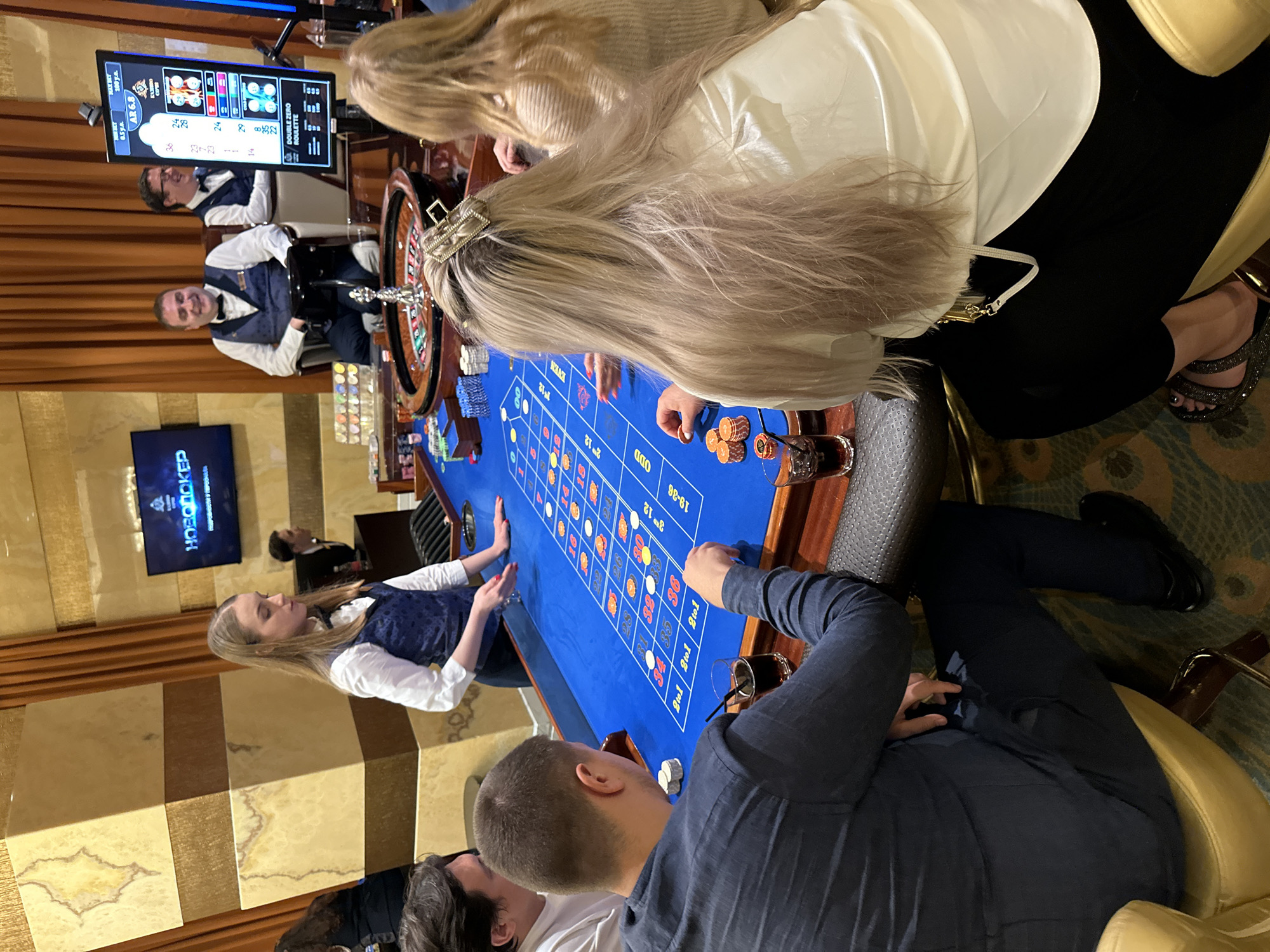 This image depicts a scene inside a casino in Estosadok, Russia, likely at a roulette table. A female croupier in a blue vest and white shirt is actively managing the game, with her hands positioned over the roulette layout. She is facing the table and appears focused on the game. On the left side of the frame, a man wearing a dark blue long-sleeved shirt and dark pants is seated at the table, with his left arm resting on the edge. He is looking towards the table, and a glass of dark liquid is placed in front of him. To his left, partially visible, is another man in a white shirt.
To the right of the croupier, two men in similar attire to the croupier are seated behind the table, observing the game. One of them, wearing a bow tie and vest, is smiling and looking towards the roulette wheel. Behind them, a large screen displays "НОВОЕ POKER" and other graphics. In the foreground, a woman with long blonde hair, wearing a white top and black pants, is seated to the right of the image. She is also looking towards the roulette table, and a small pile of orange casino chips is in front of her. Another woman with blonde hair, wearing a beige sweater, is seated further back on the right, also looking towards the table.
The environment appears to be a well-lit casino interior with ornate wall coverings featuring gold and beige marble-like patterns and brown curtains. The roulette table is covered in a vibrant blue felt with a detailed betting layout. Several stacks of chips are visible on the table. The overall atmosphere suggests an active gaming session.