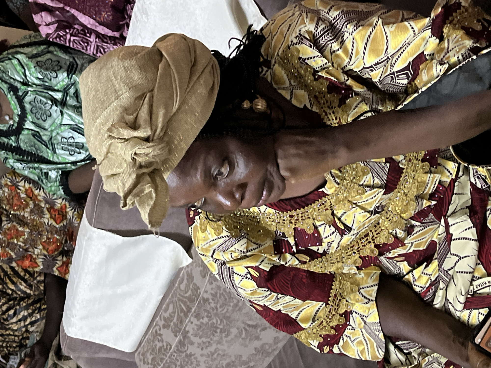 A woman is pictured from the chest up, sitting on a sofa in Du, Nigeria. She is wearing a gold headwrap and a yellow, maroon, and gold patterned dress. Her left hand is resting on her chin as she looks to her right. Other people are visible in the background, also seated and wearing colorful traditional clothing.