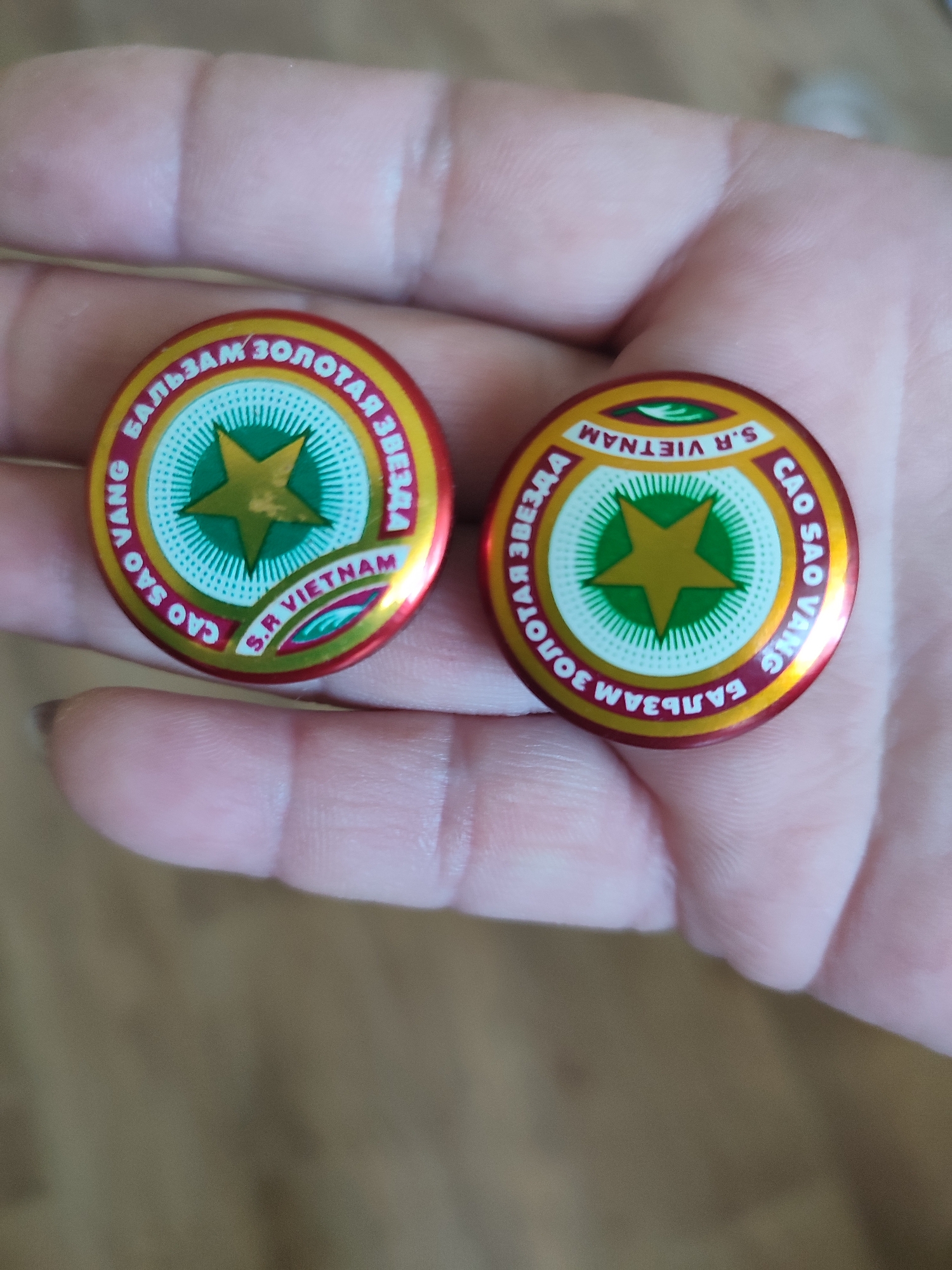 This media file shows a close-up of two round, metallic badges held in a person's hand. The badges are red with yellow rims and feature a central green star on a white, radially patterned background.
The badge on the left has text around the circumference. Partially visible text includes "БАЛЬЗАМ ЗОЛОТАЯ ЗВЕЗДА" in Cyrillic and "CAO SAO VANG S.R VIETNAM" in Latin script. The badge on the right also has text around the circumference, appearing to be the same as the left badge but oriented differently. The text in Latin script "S.R VIETNAM" is clearly visible on the right badge.
The person's skin is visible, with fingers gently holding the badges. The background is blurred, suggesting a shallow depth of field, and appears to be a neutral, earthy tone. The lighting is soft, indicating an indoor setting or an overcast day. The overall impression is that of a casual presentation of the objects. No specific event or emotional context is conveyed beyond the presentation of the items. The location context provided is Narowlya, Belarus, which might be relevant to the origin or context of the badges.