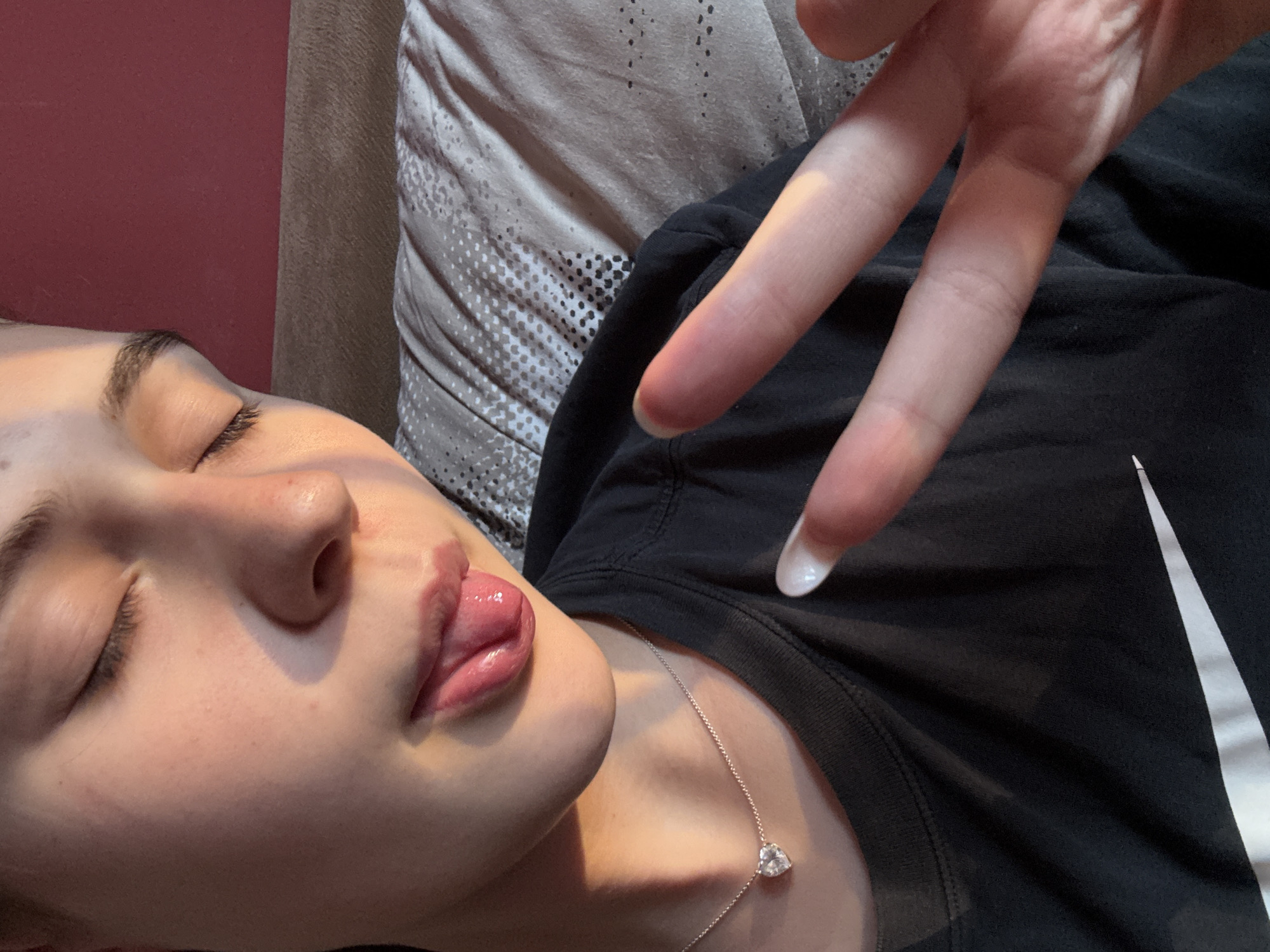 A close-up shot shows a person's face and upper torso. The person's eyes are closed, and their tongue is sticking out. They are wearing a black t-shirt and a silver necklace with a heart-shaped pendant. Their right hand is raised, with two fingers extended in a peace sign. The background is a blurred image of a bed and a reddish wall. The lighting suggests it is either sunrise or sunset, casting a warm glow on the person's face. There is no discernible information about the location in Panagyurishte, Bulgaria.