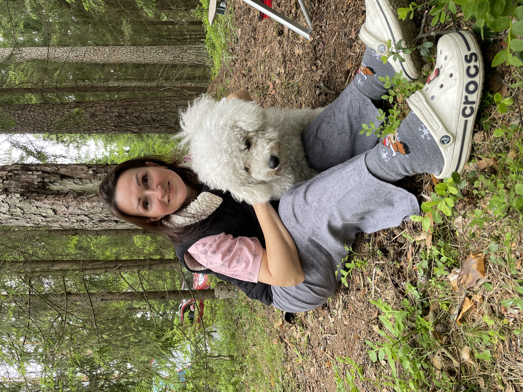 A middle-aged woman of European descent with dark brown hair is sitting on the ground in a forest, holding a fluffy white Bichon Frise dog. She is wearing a pink t-shirt with a squirrel pattern, a black vest with a sherpa collar, grey pants, and white Crocs with festive socks. The dog is looking to its right, away from the camera. The background consists of tall pine trees and green undergrowth. The ground is covered with pine needles and fallen leaves. The overall scene appears to be a relaxed outdoor setting, possibly in or around Roshchino, Russia, given the dense forest environment.