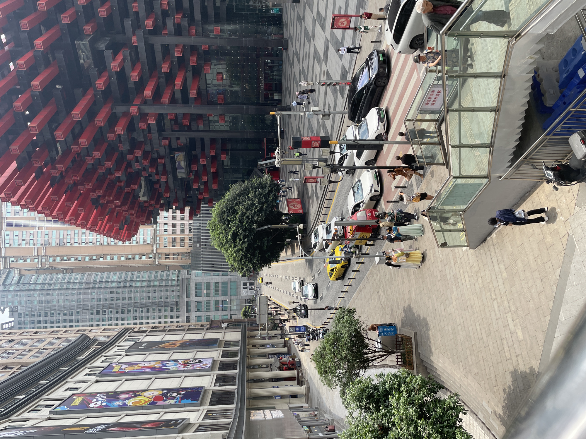 This is an eye-level, outdoor photograph of a street in Yuzhong District, Chongqing, China. The view is from an elevated position, looking down the street. Pedestrians, cars and trees line the street. On the left, there is a building with many large posters on its façade. Adjacent to this building, and extending down the street, are buildings of a typical city design. A peculiar, modern building with red rectangular accents stands out on the upper left of the image. At the top, there is a pedestrian bridge with glass railings. Several people are visible on the street and crosswalks. There is normal city traffic including cars, trucks, and taxi cabs.