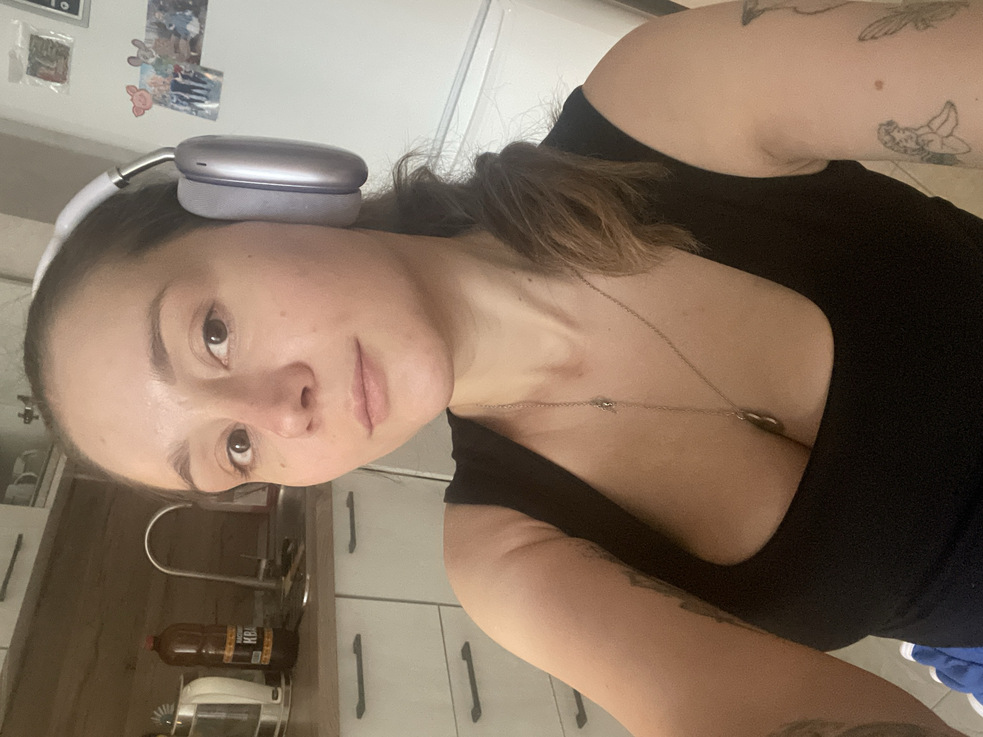 The image features a close-up, head-and-shoulders shot of a person wearing large, silver headphones. The person is looking directly at the camera with a neutral expression. They are wearing a black tank top, and a delicate silver necklace is visible around their neck. The background appears to be a kitchen, with wooden cabinets and drawers visible. There is a bottle of what appears to be a beverage with Cyrillic text on it. In the upper left corner, there are some small framed pictures or photos on the wall. The lighting suggests it could be daytime. The person has tattoos on their arm.