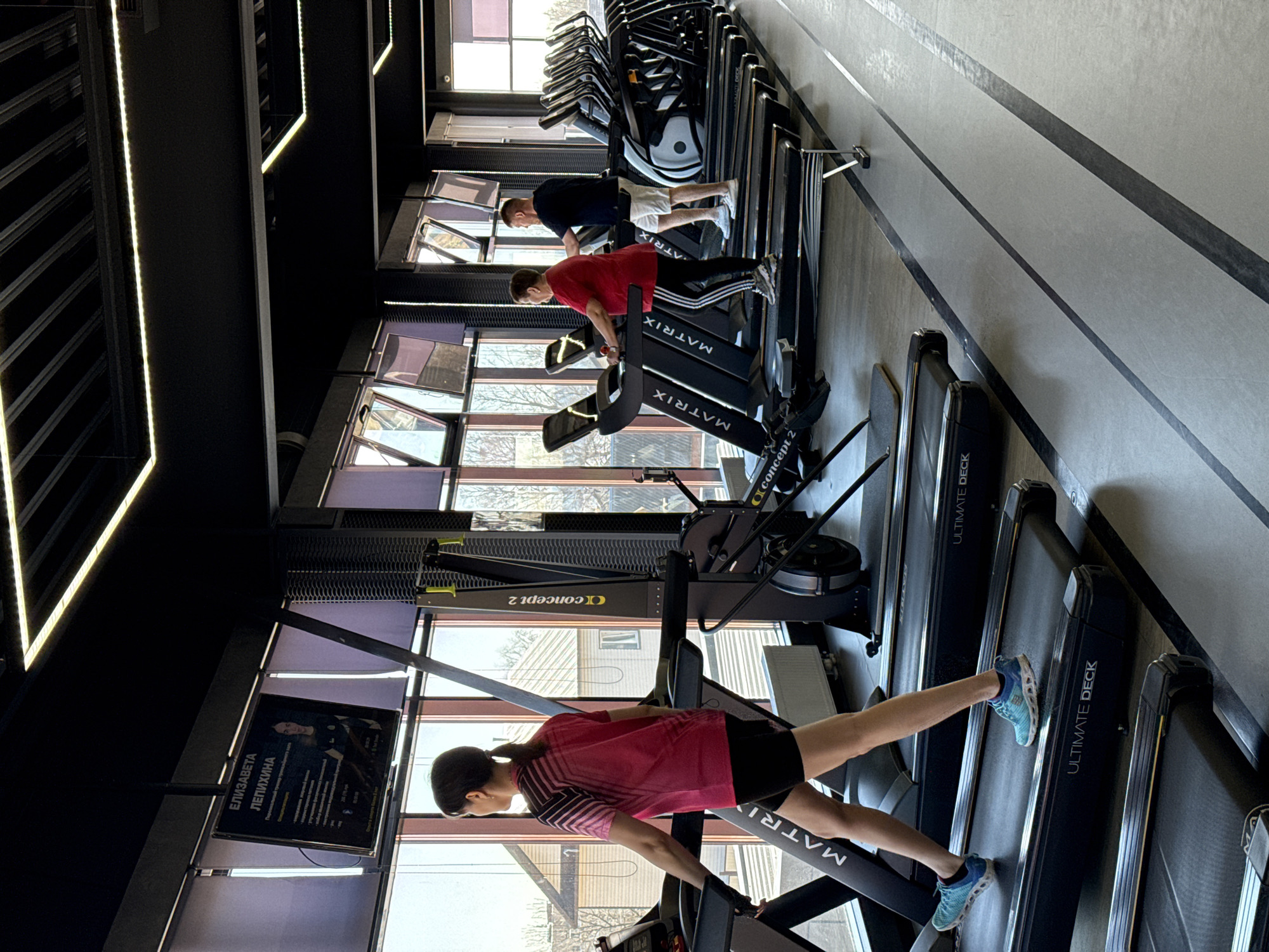 A gym in Votkinsk, Russia, features multiple treadmills and rowing machines. Three people are visible, two are using treadmills, and another is further back. A woman in a pink shirt is using a treadmill closer to the camera. A screen displays information about Elizaveta Lepikhina. The gym has large windows and modern lighting.