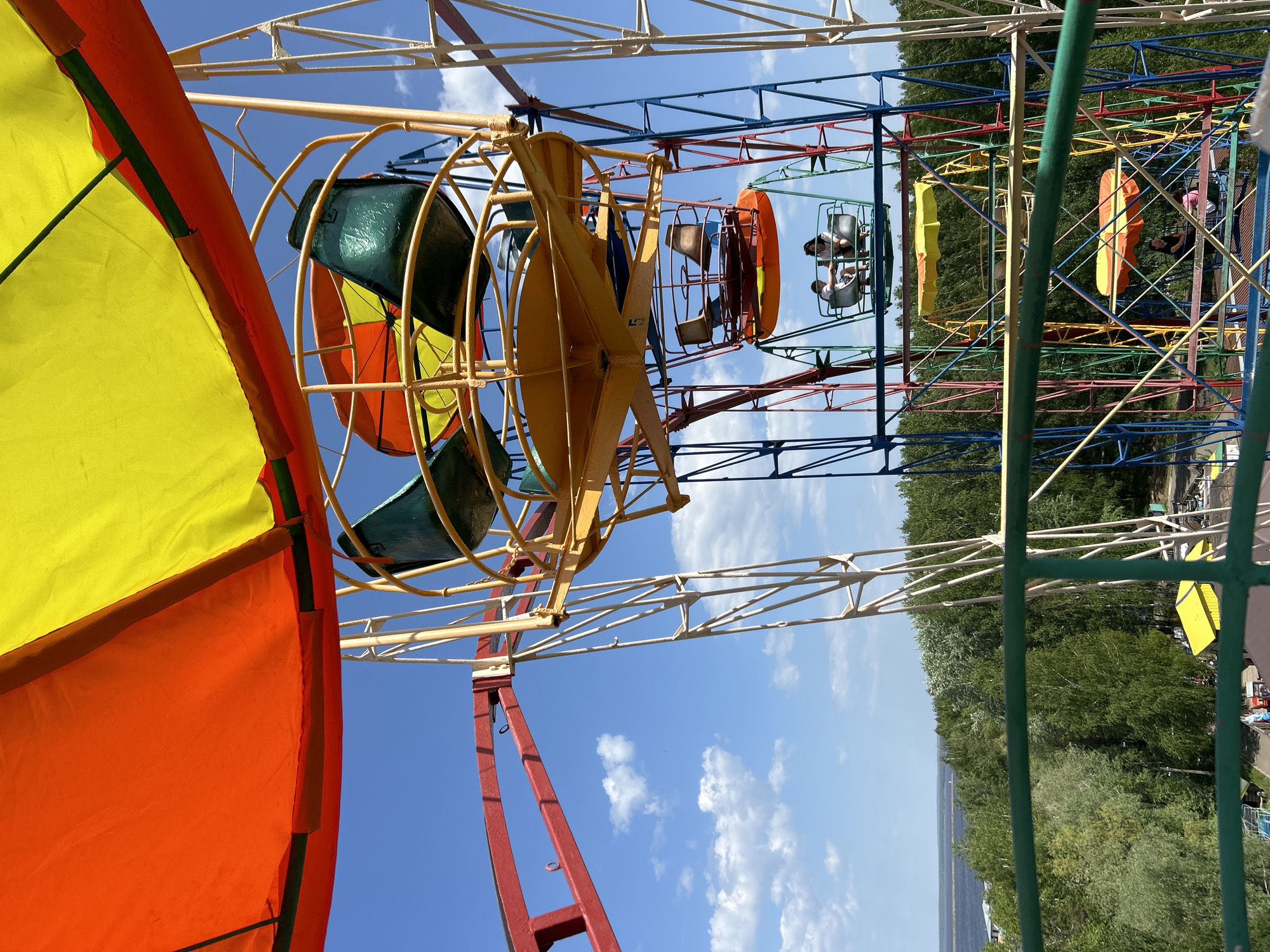 The image shows two carnival rides against a backdrop of a blue sky with scattered clouds, a treeline, and a body of water.
One ride features enclosed capsules, some occupied by individuals who appear to be sitting. The framework supporting the capsules is a brightly colored array of red, blue, yellow, and green.
The other ride has a more open design with rotating circular structures. These structures consist of frames with curved sections of varying colors, including yellow, green, and orange.
The setting is likely a park or fairground in the city of Chaikovsky, Russia, given the presence of typical amusement park rides and natural background elements.