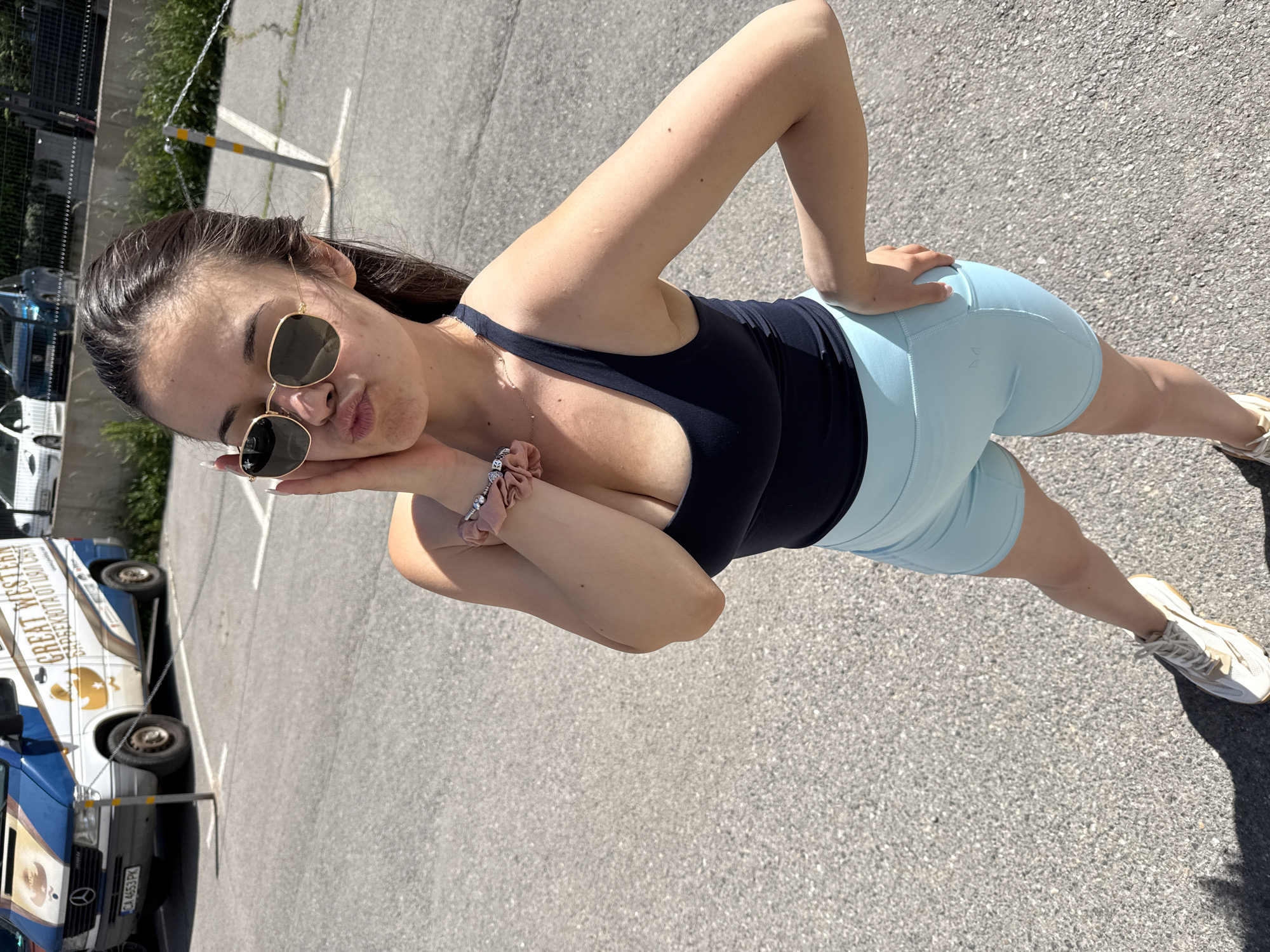 A young woman is standing outdoors in an asphalt parking lot or paved area. She is wearing a dark sports bra or tank top and light blue biker shorts. She has on sunglasses and a scrunchie around her wrist. She's striking a pose with one hand on her hip and the other near her face, puckering her lips. In the background, there is a white van with the words "GREAT WESTERN" written on it. The van has a license plate that reads "CA 4653 PK." There are buildings and some greenery also visible in the background. It's daytime and sunny. Based on the license plate number CA4653PK, this parking lot is located in Sofia, Bulgaria.