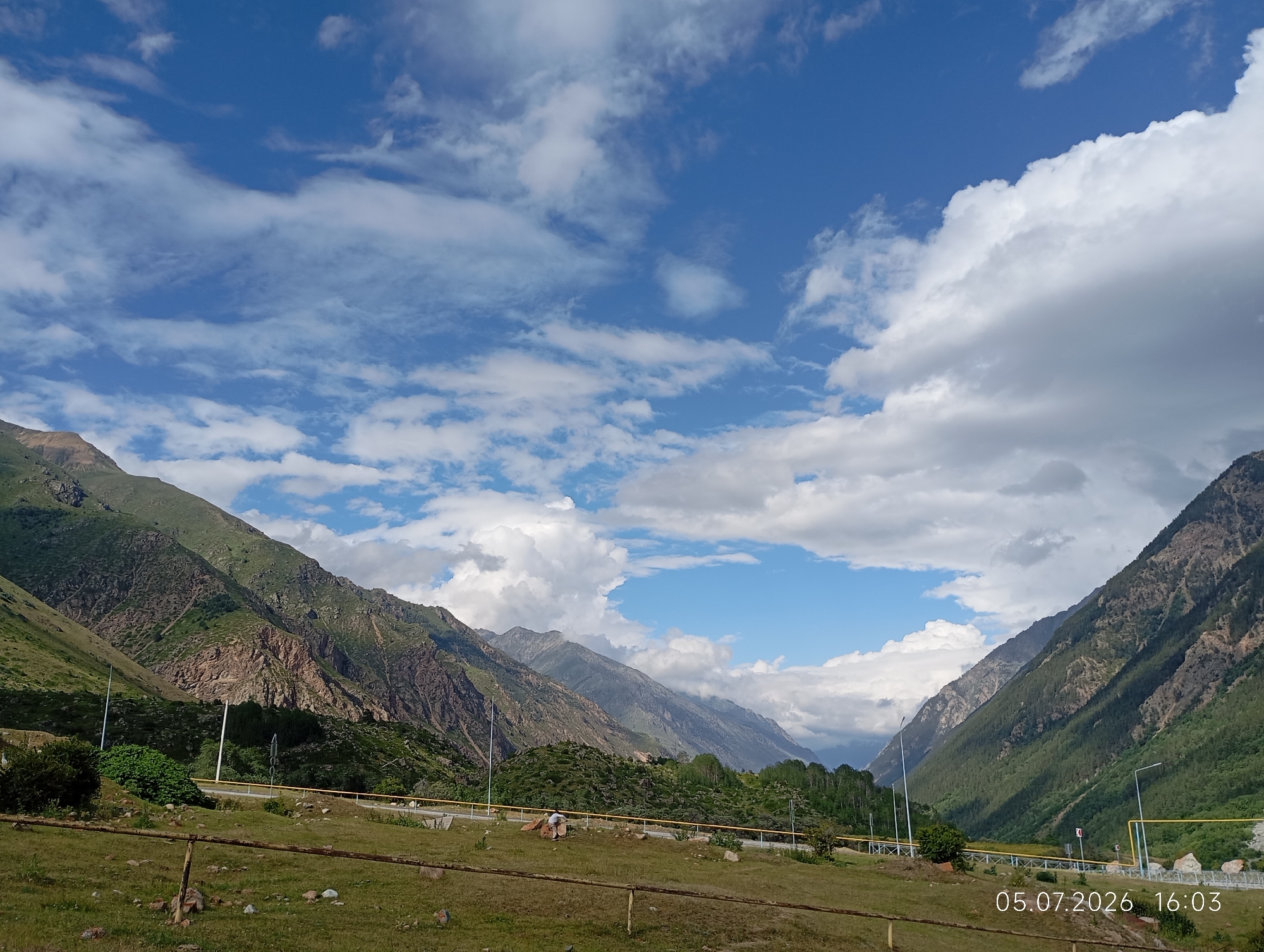 This is a landscape photograph taken in the city of Neutrino, Russia. The image captures a valley surrounded by steep, green mountains under a bright blue sky filled with white, fluffy clouds. A paved road runs through the valley, bordered by a yellow railing and lampposts. In the foreground, a grassy field is visible, with a wooden fence and some scattered rocks. The date stamp on the image indicates it was taken on July 5, 2026, at 16:03.