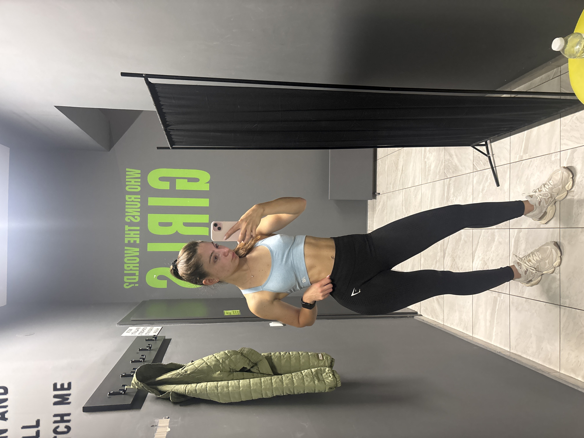 A young woman is taking a mirror selfie in what appears to be a gym locker room. She is wearing a light blue sports bra and black leggings, with white athletic shoes. A green puffer jacket hangs on a coat rack to her left. The walls are dark gray, with a large, bright green graphic on the wall behind her that reads "WHO RUNS THE WORLD? GIRLS." To her right, a black curtain hangs from a frame. The floor is tiled with a light gray and white pattern. A yellow object, possibly a bench or table, is visible on the far right, with a water bottle on top. Text is visible on the wall to the left, part of which reads "WATCH ME." The image is taken in Varna, Bulgaria.
