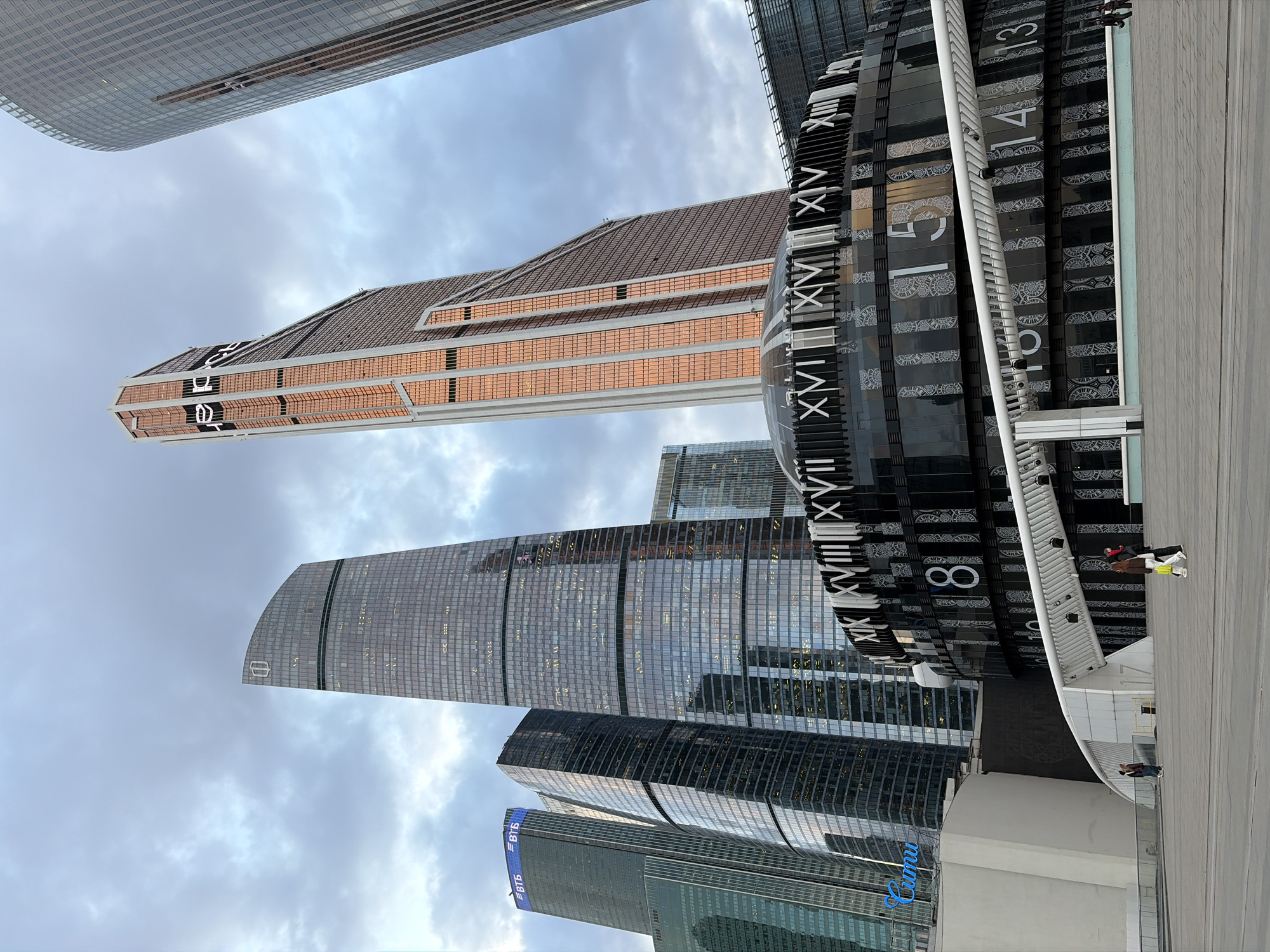 The image captures an urban landscape in Khimki, Russia, featuring a cluster of modern skyscrapers against a cloudy sky. Dominating the foreground is a large, decorative clock structure with Roman numerals and number 15. In the immediate foreground is a flat, paved surface with a few individuals walking. The buildings are of various heights and architectural designs, with glass exteriors reflecting the sky. Signage on one of the lower buildings features a "Crocus" logo. The overall composition suggests a commercial or business district within the city.