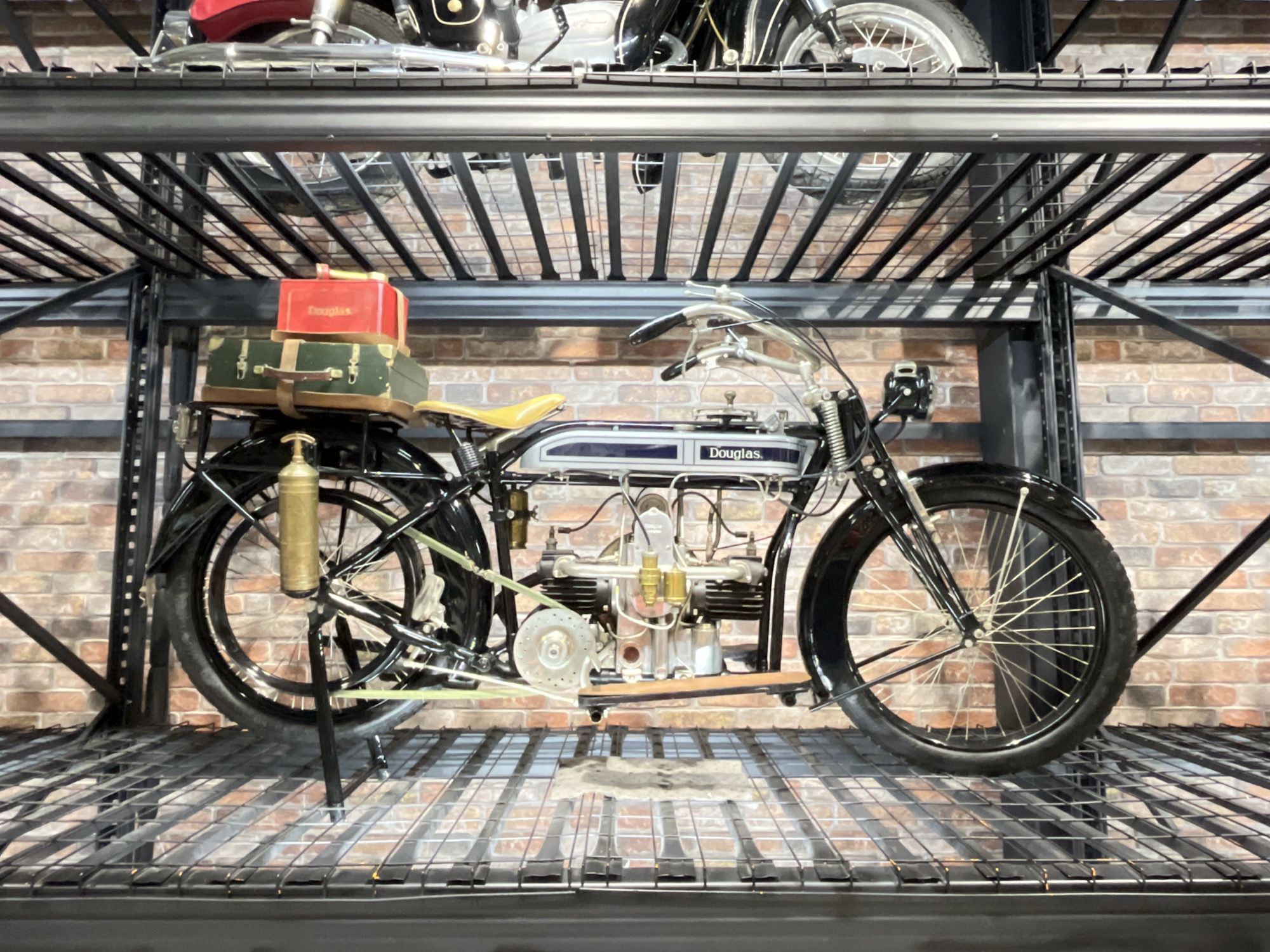 The image shows a vintage Douglas motorcycle displayed on a black metal shelf. The motorcycle is mostly black with a blue and white fuel tank and a yellow seat. It features a large engine with visible cylinders and exposed mechanics. On the rear rack, there are stacked suitcases, one green and the other red. The motorcycle appears to be restored and is in a static display setting. The backdrop is a brick wall. The display is on a rack with another motorcycle above it.